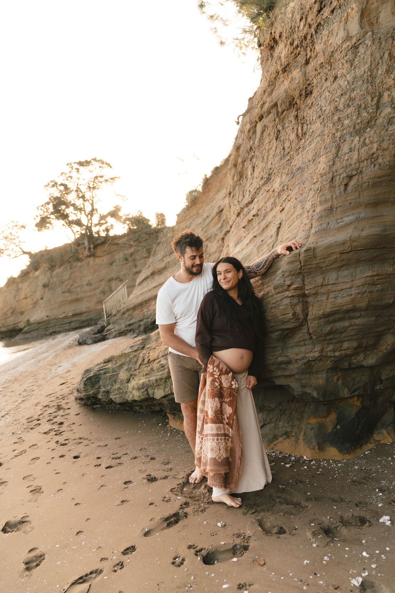 A couple posing for maternity photos against rocky cliffs and sand at sunset on a beach.