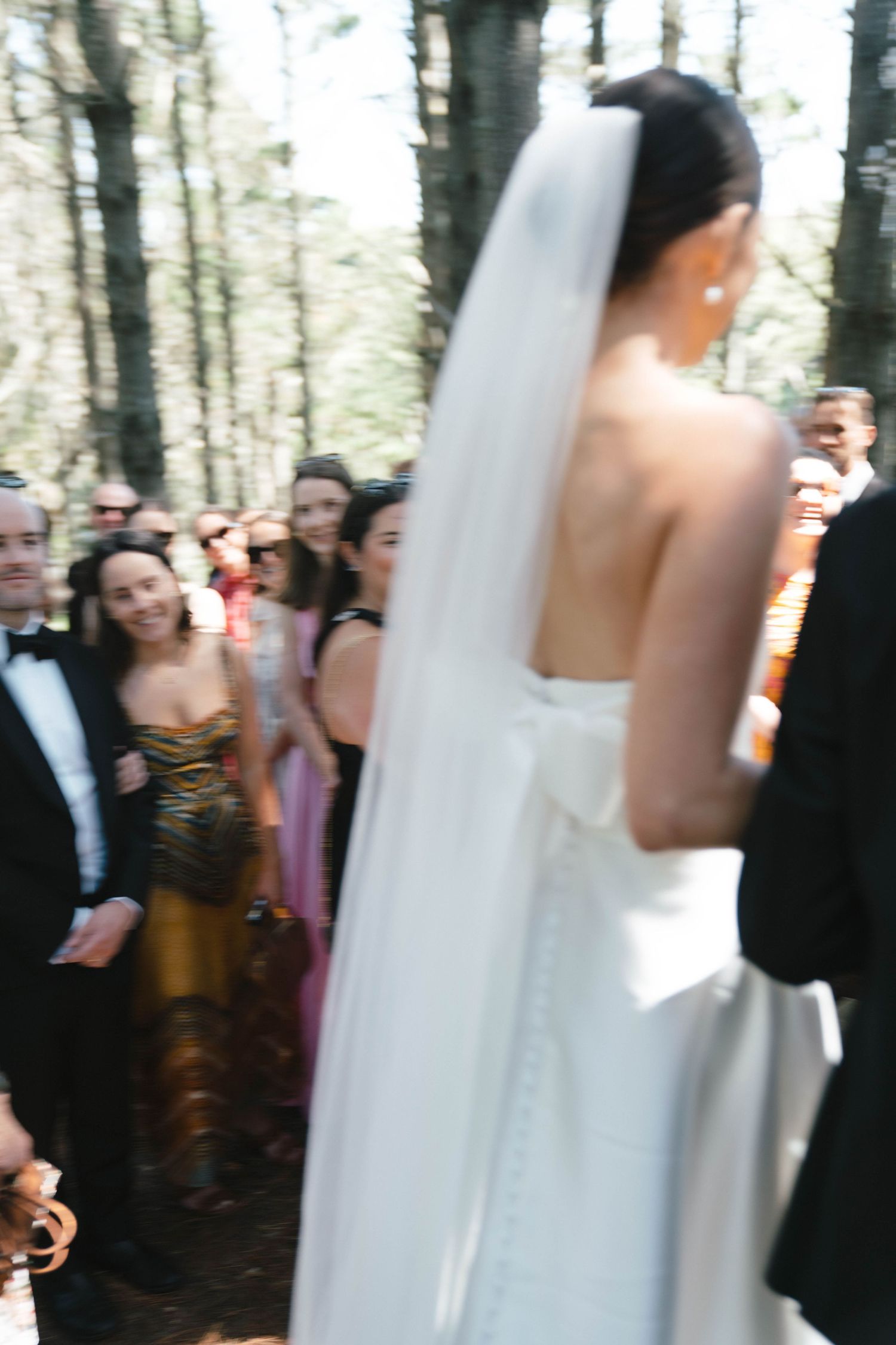 Wedding guests watch as the ceremony unfolds in a wooded setting.