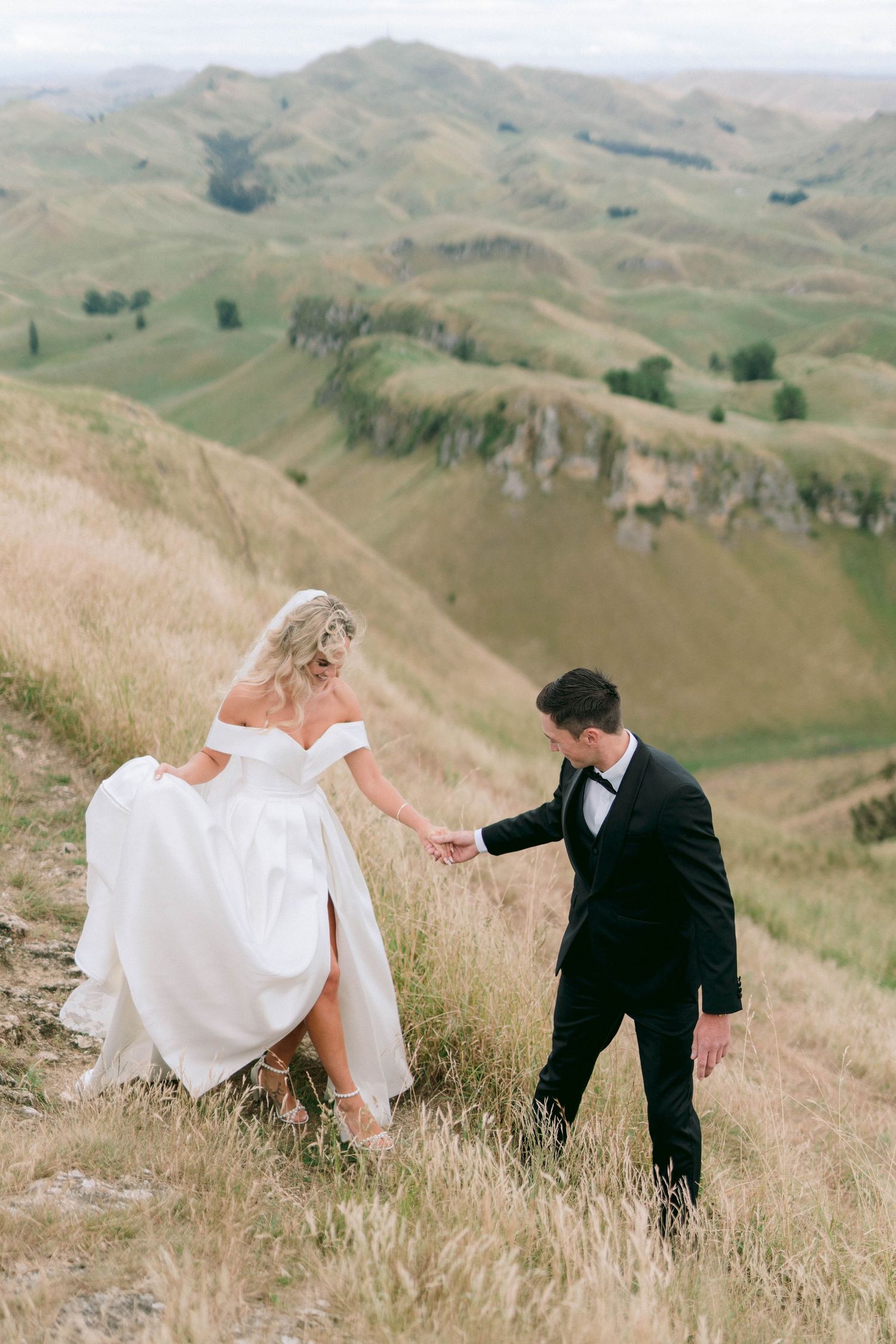 A wedding couple walking along a scenic hilltop path with rolling hills in the background.