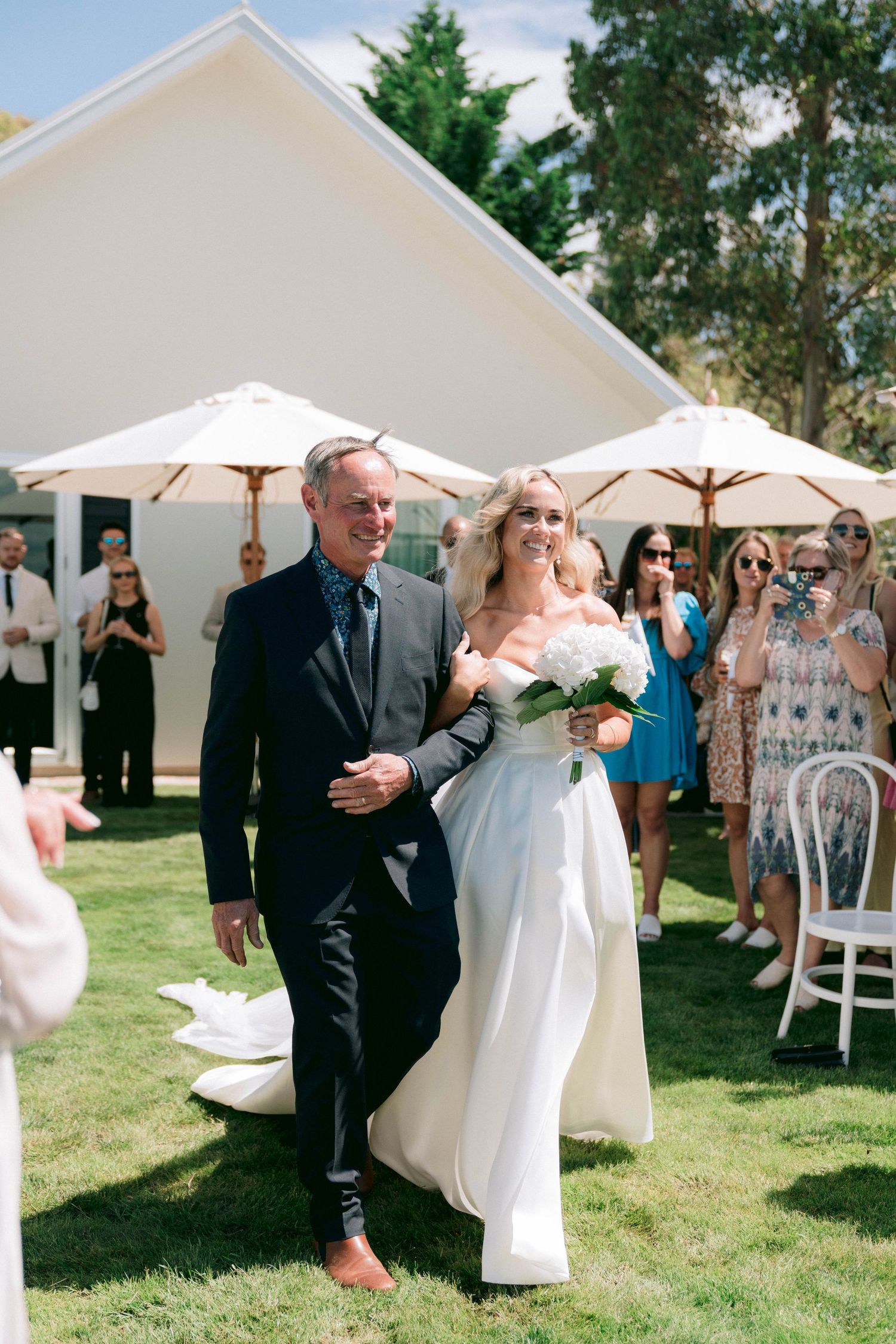 Outdoor wedding ceremony processional under white umbrellas on a sunny lawn.