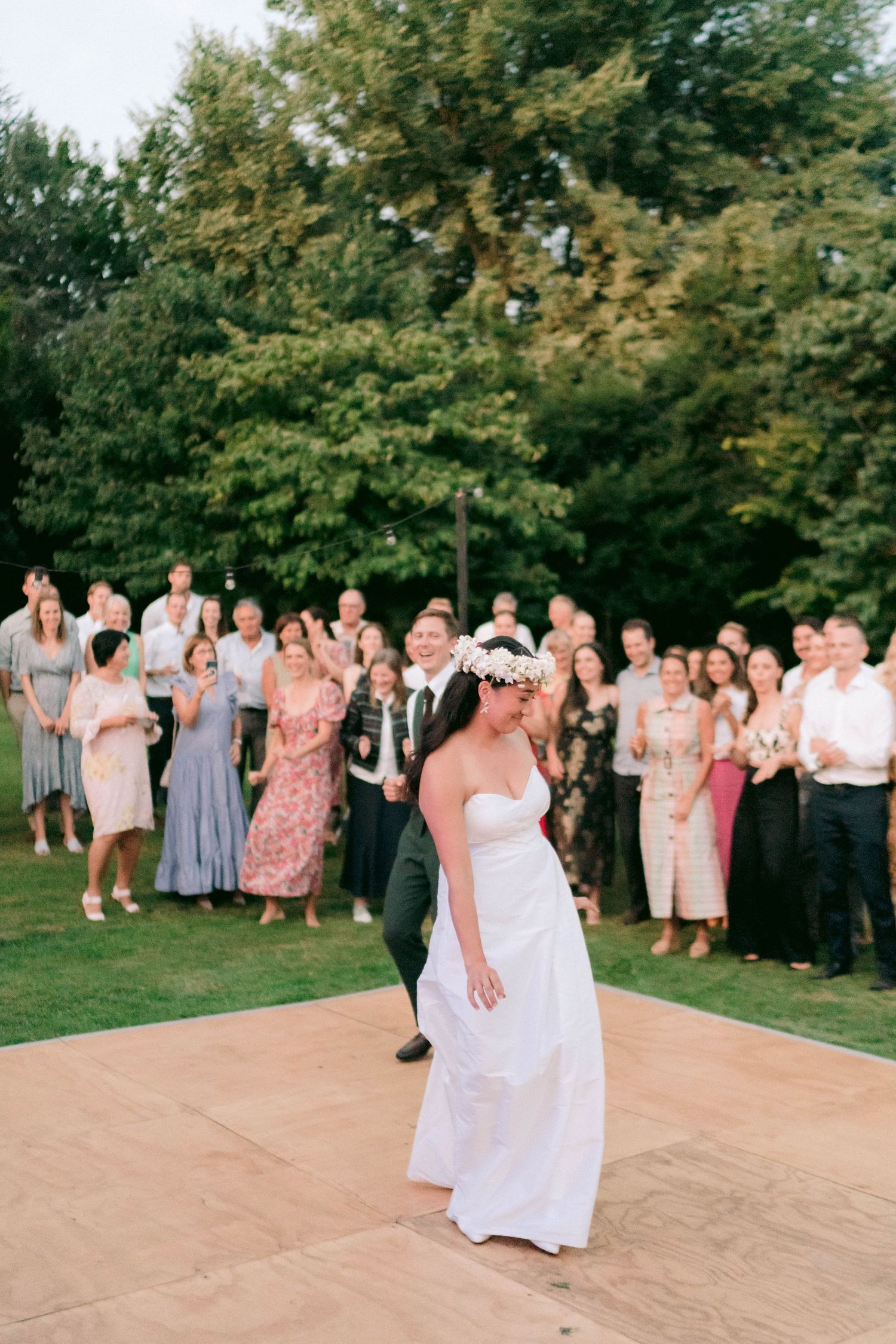 A wedding couple dancing on an outdoor dance floor with guests watching in the background during sunset.