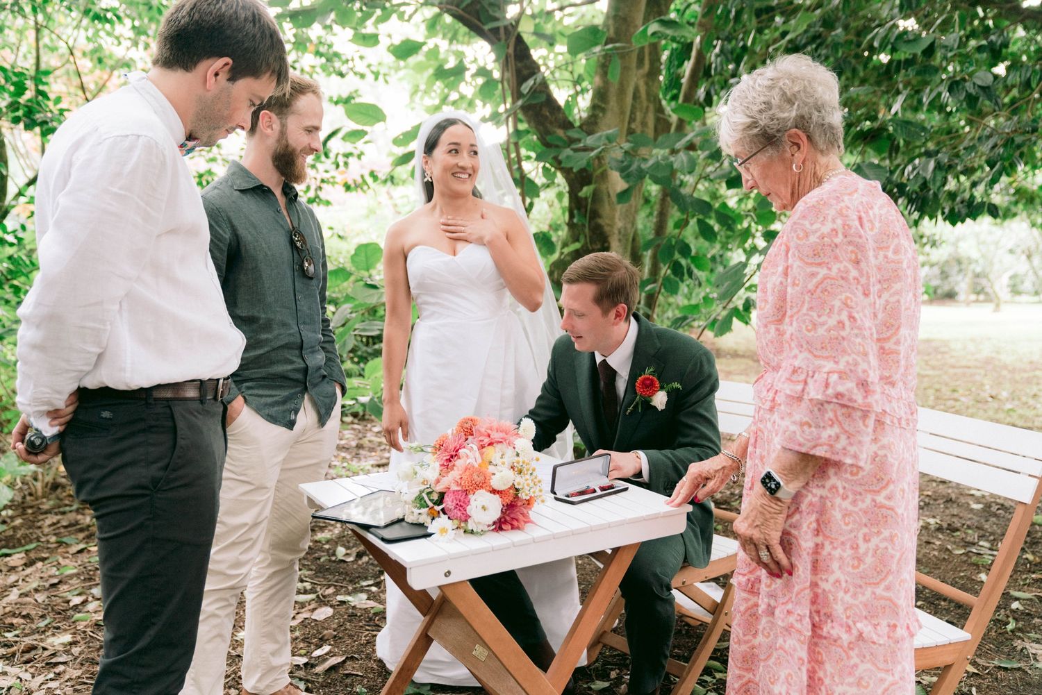 Wedding party gathered around a table signing official documents outdoors.