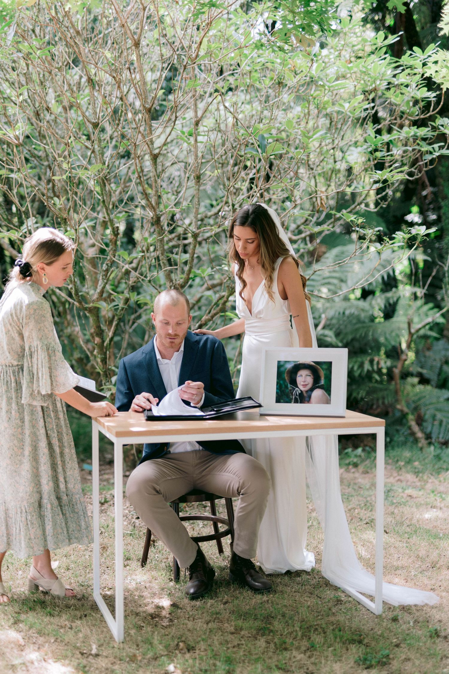 People signing documents at a white table outdoors in a garden setting for a special occasion.