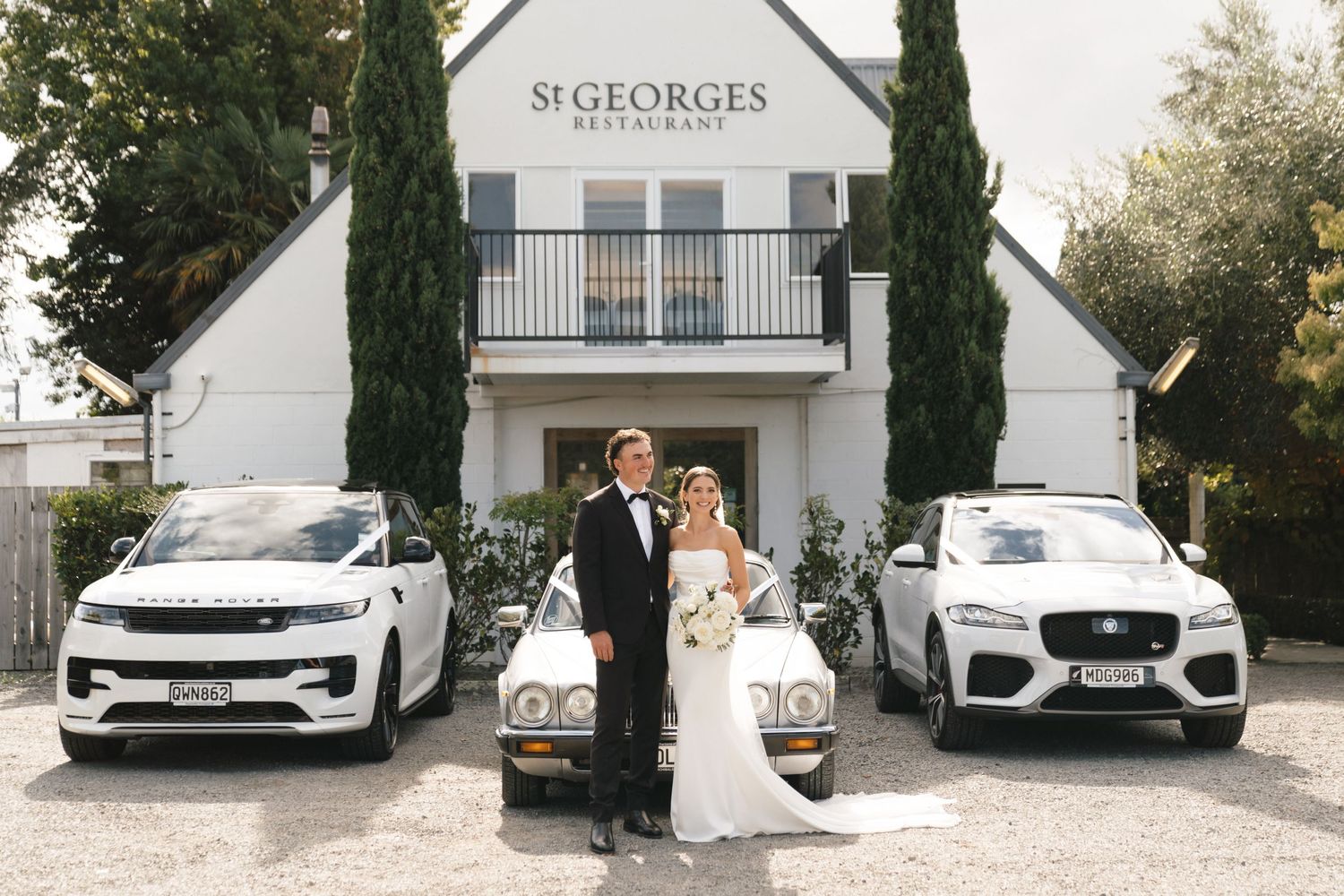 Luxury white cars parked outside an upscale venue with cypress trees framing the entrance.