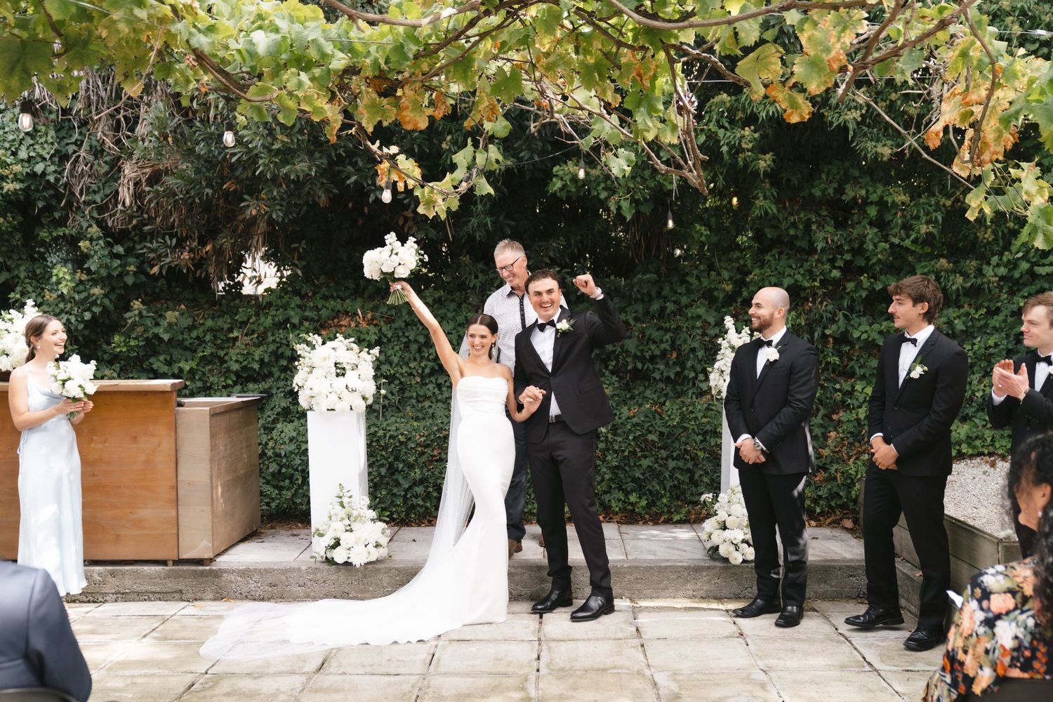 Outdoor wedding ceremony setup with guests in formal attire under trees.