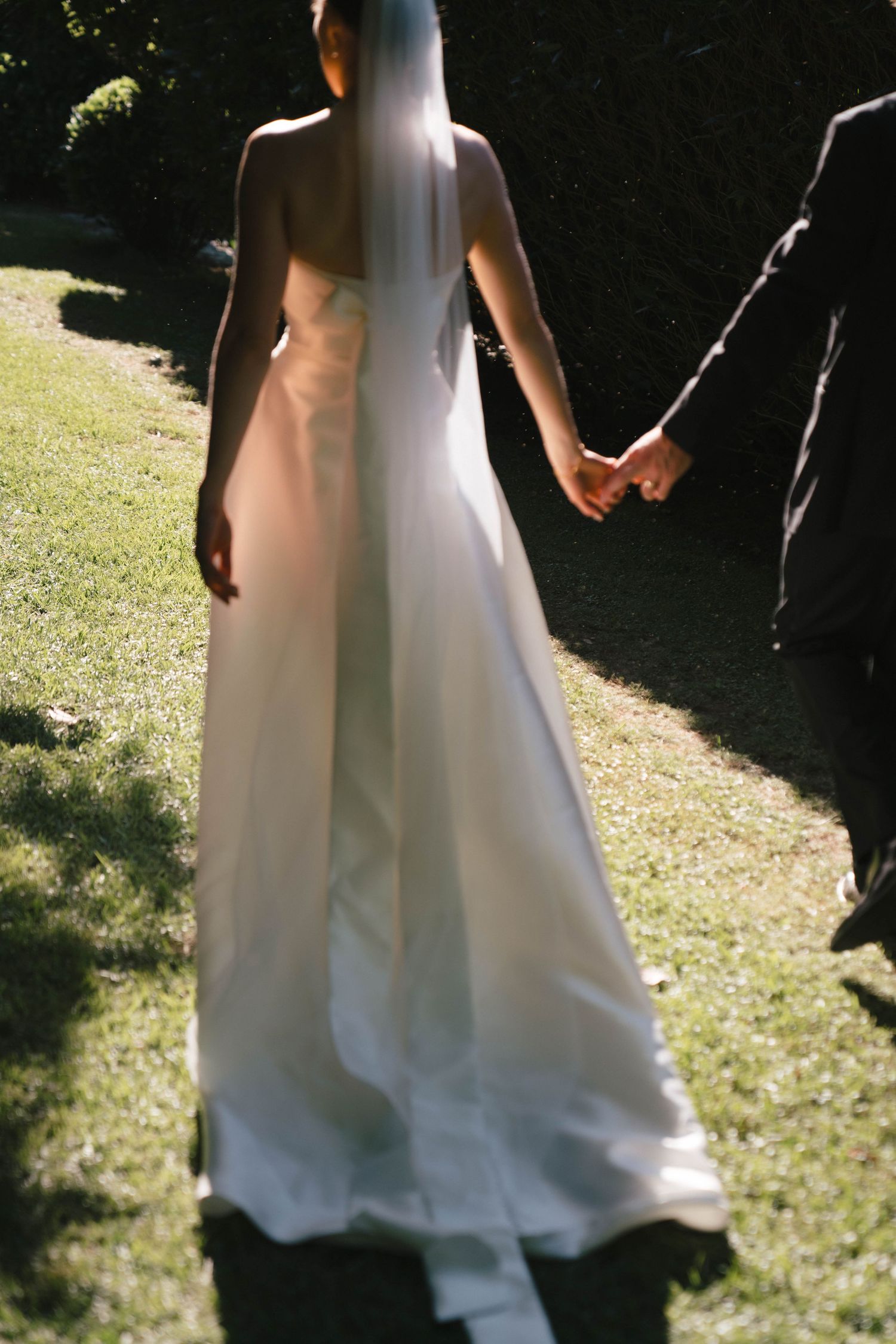 Elegant white wedding dress flowing in sunlight as couple holds hands in a garden setting.