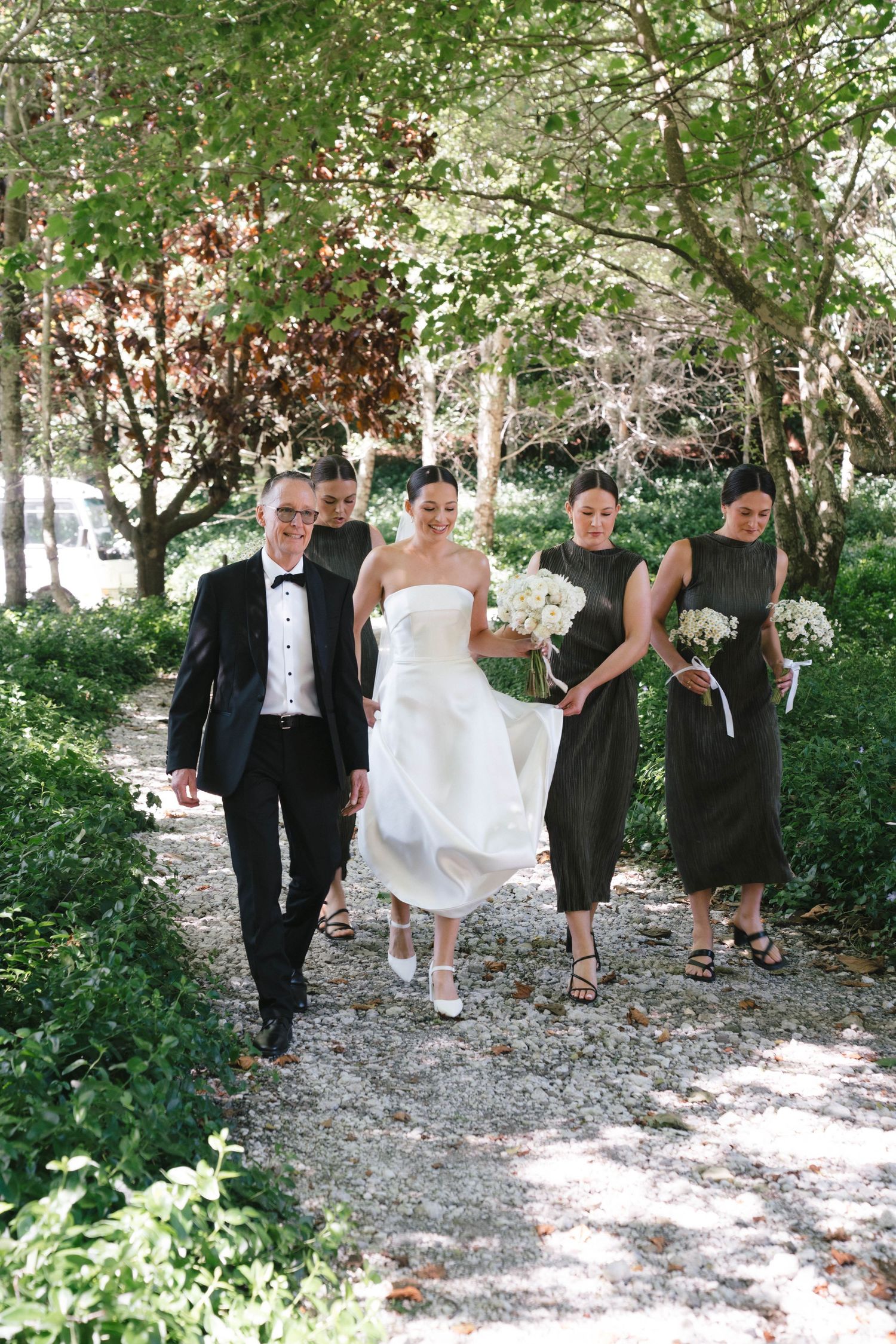 Wedding party walks down a garden path with attendants in dark dresses.