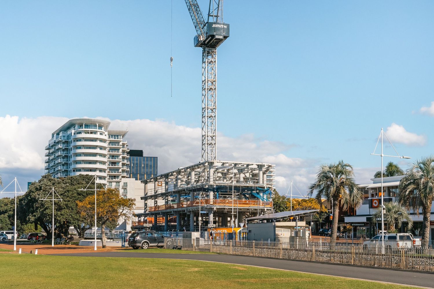 Modern construction project with tower crane and high-rise buildings visible in background near palm trees.