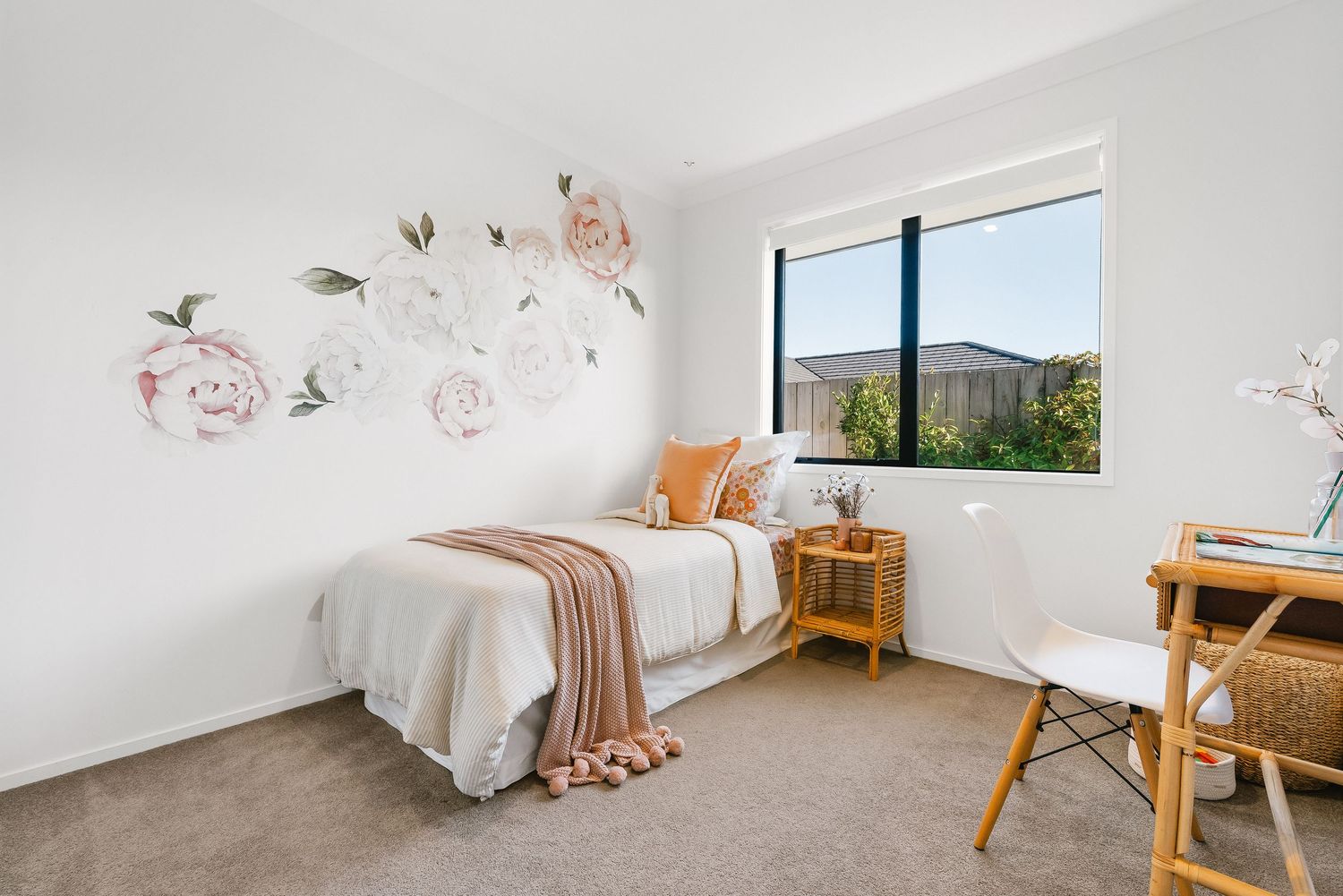 Modern bedroom with floral wall decals, white bedding, peachy accents, and a window overlooking outdoor scenery.
