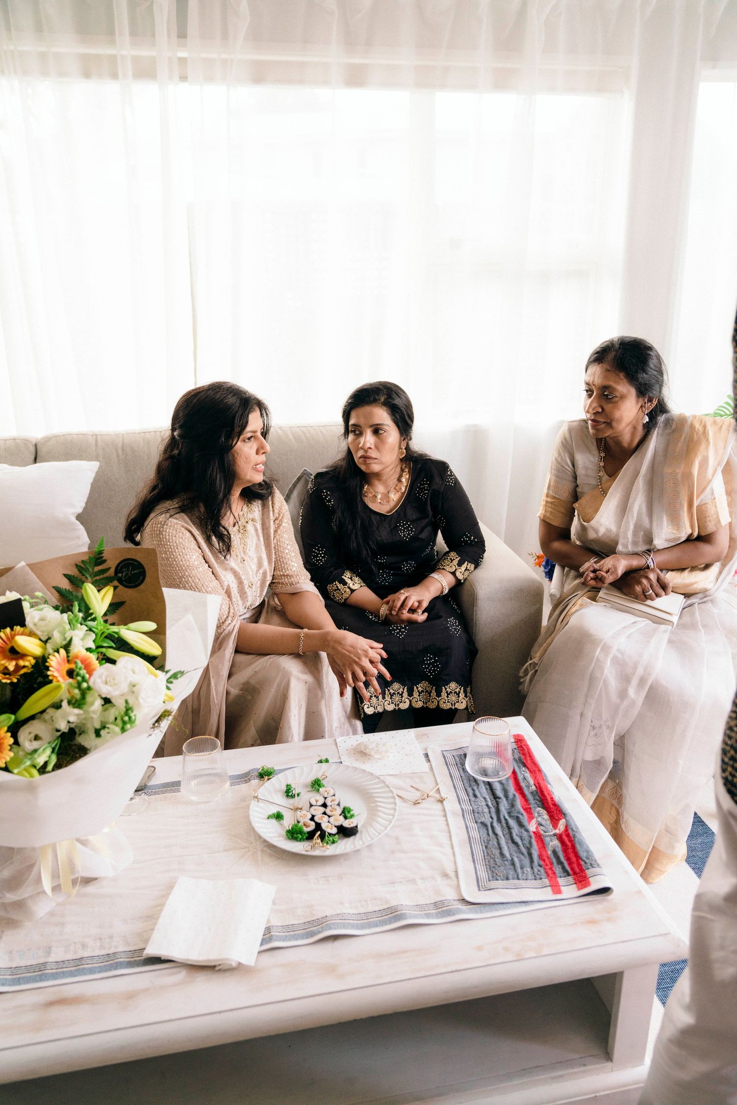 Three women in casual attire having a conversation on a couch with flowers and a coffee table in the foreground.