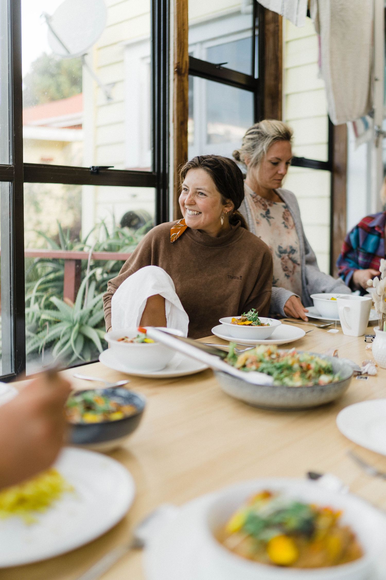 A group enjoys a sunny brunch gathering with colorful plates of food at a window-side table.