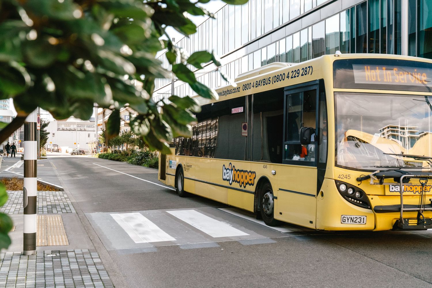 A yellow city bus waits at a crosswalk near a modern building with green foliage in the foreground.