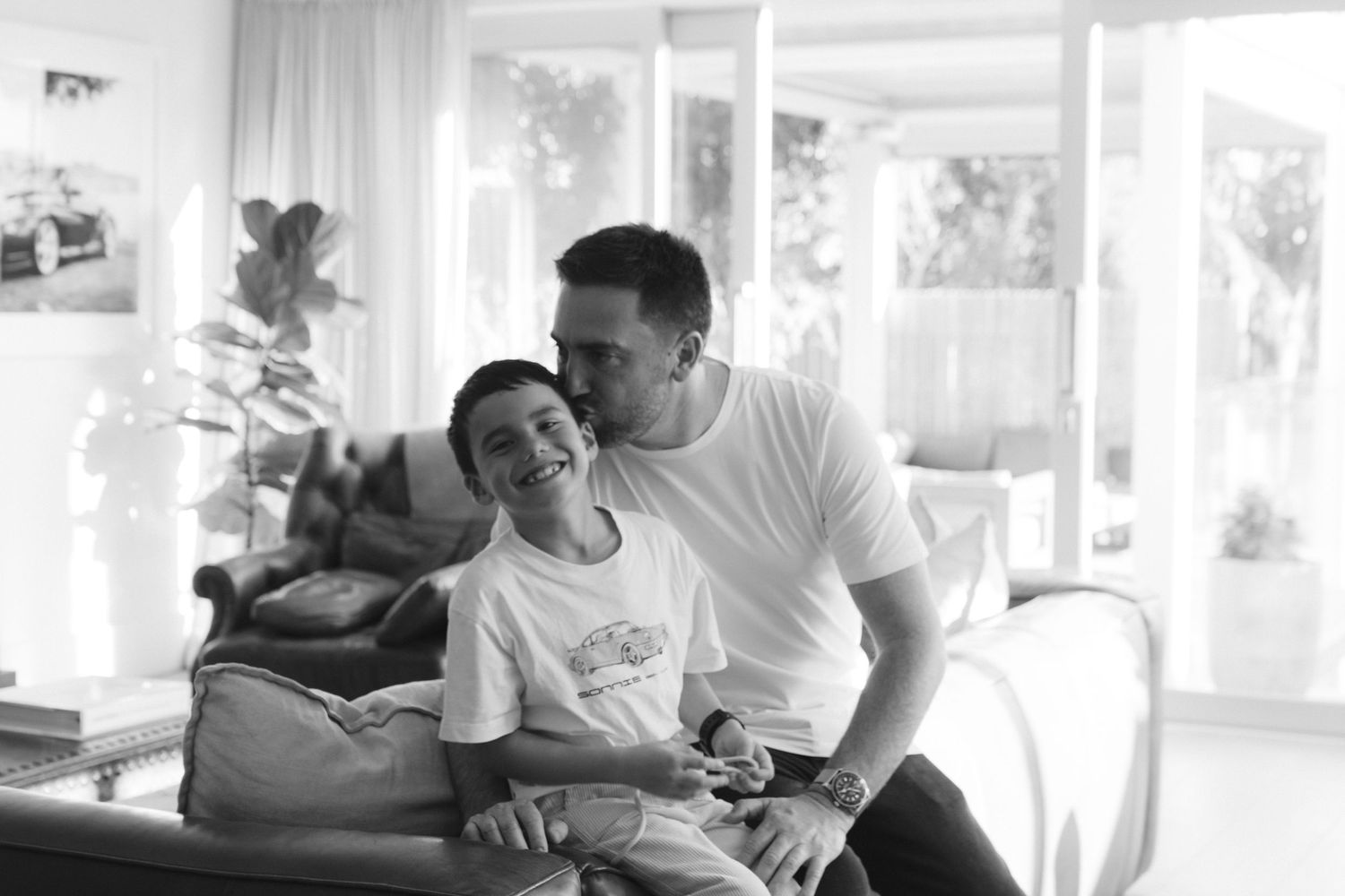 Black and white image showing an affectionate moment between parent and child sitting together on a couch in a sunny room.
