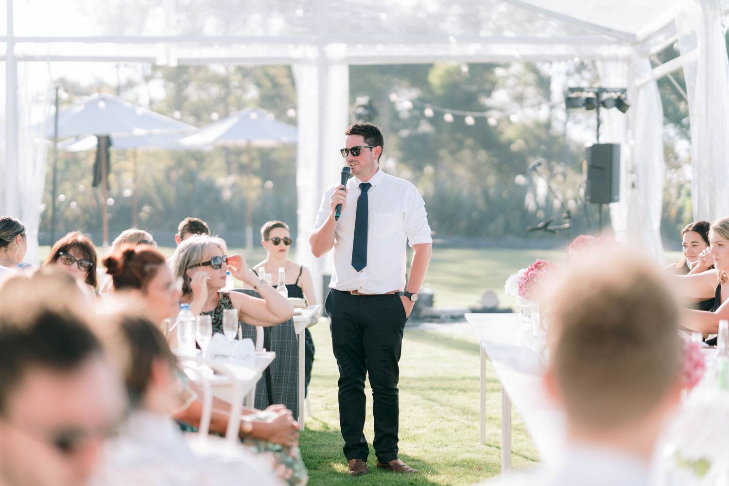 Wedding reception speech being delivered outdoors under white tent structure.