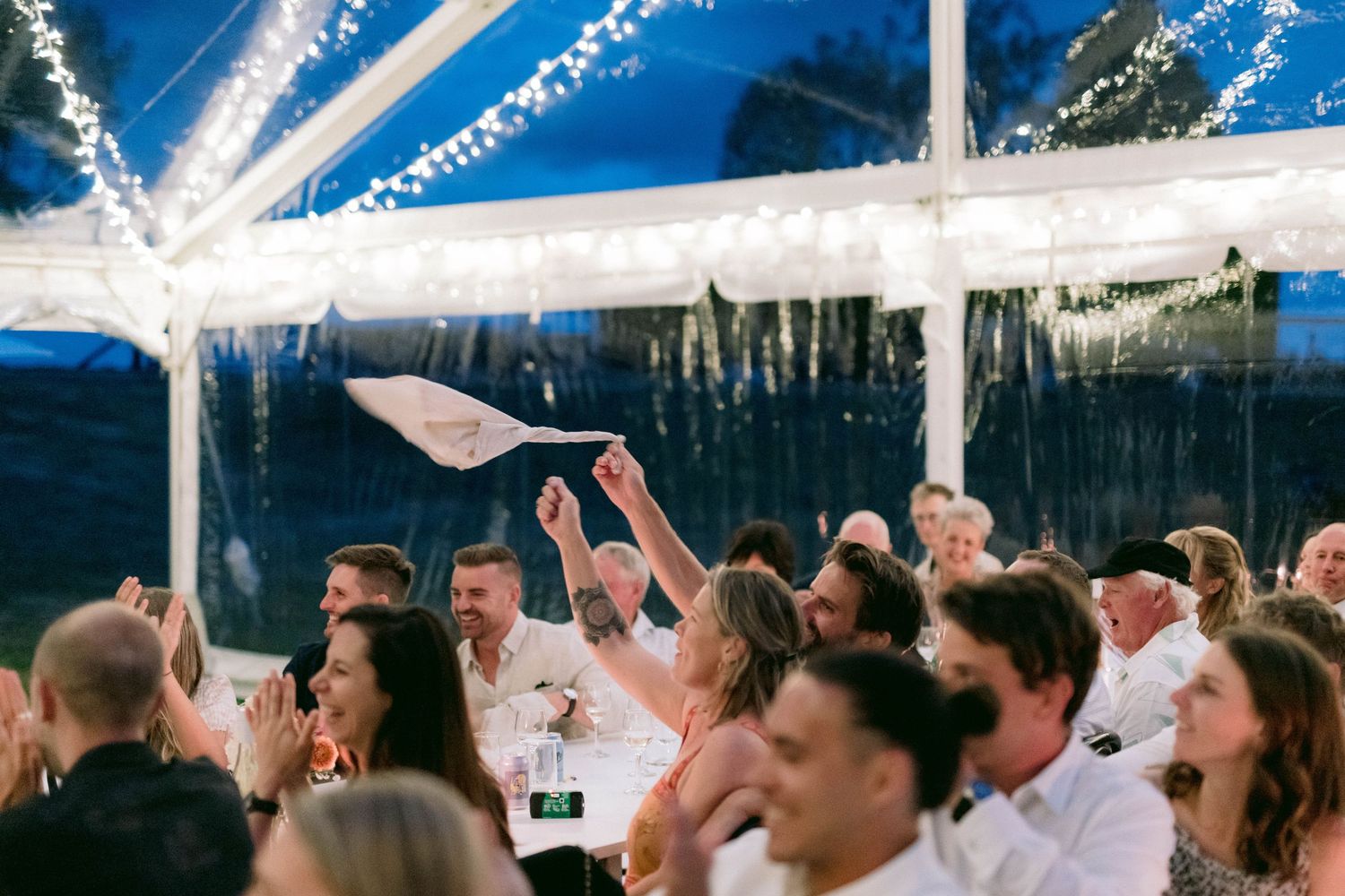 Wedding guests dance and celebrate with raised hands under a white tent at an outdoor reception.