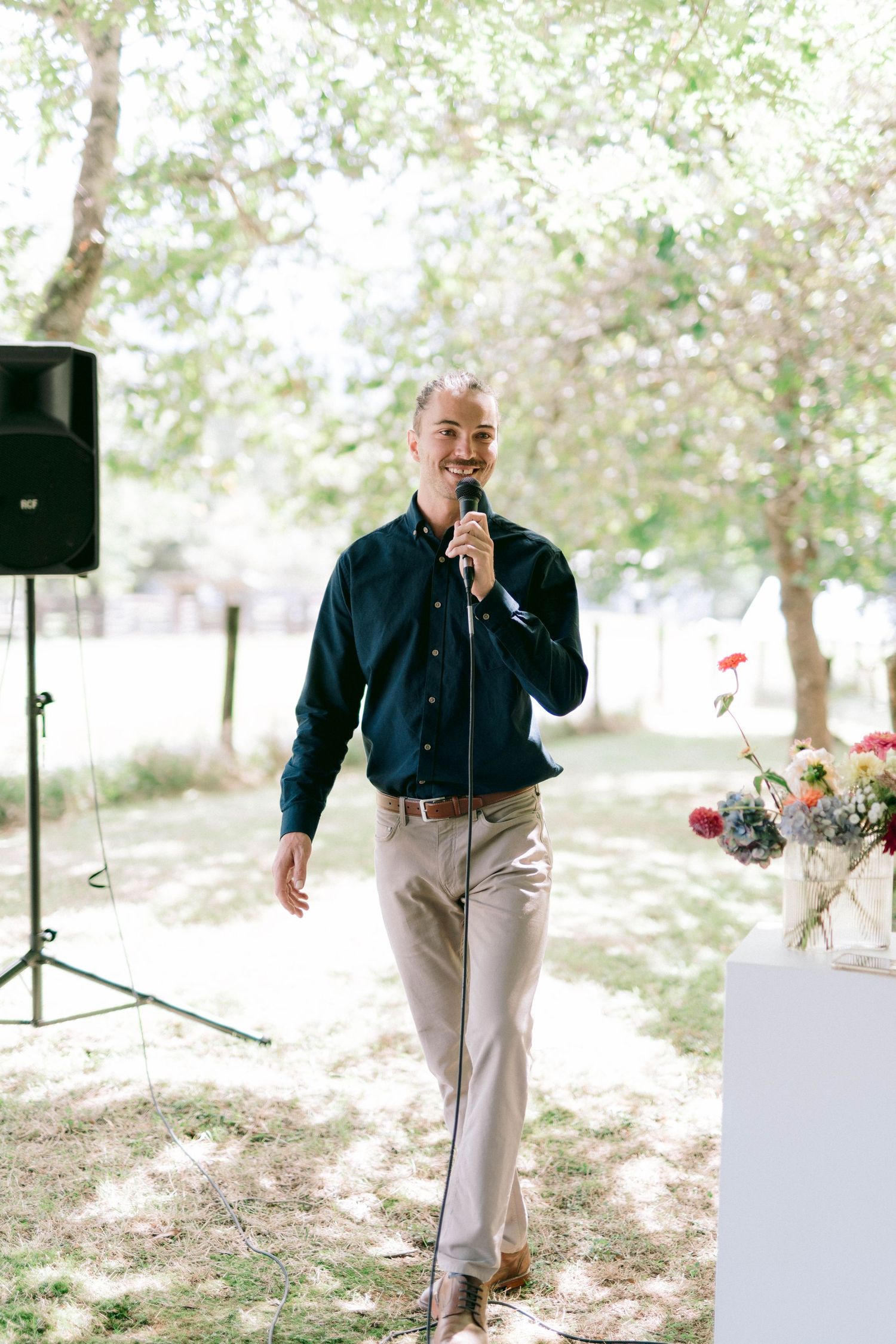 Speaker holds a microphone while giving a speech at an outdoor event with audio equipment and floral decorations.