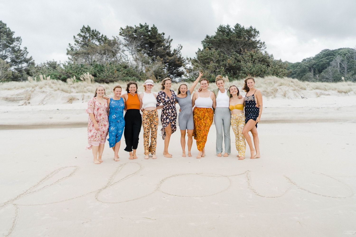 A group in coordinated summer outfits poses together on a sandy beach with pine trees in the background.