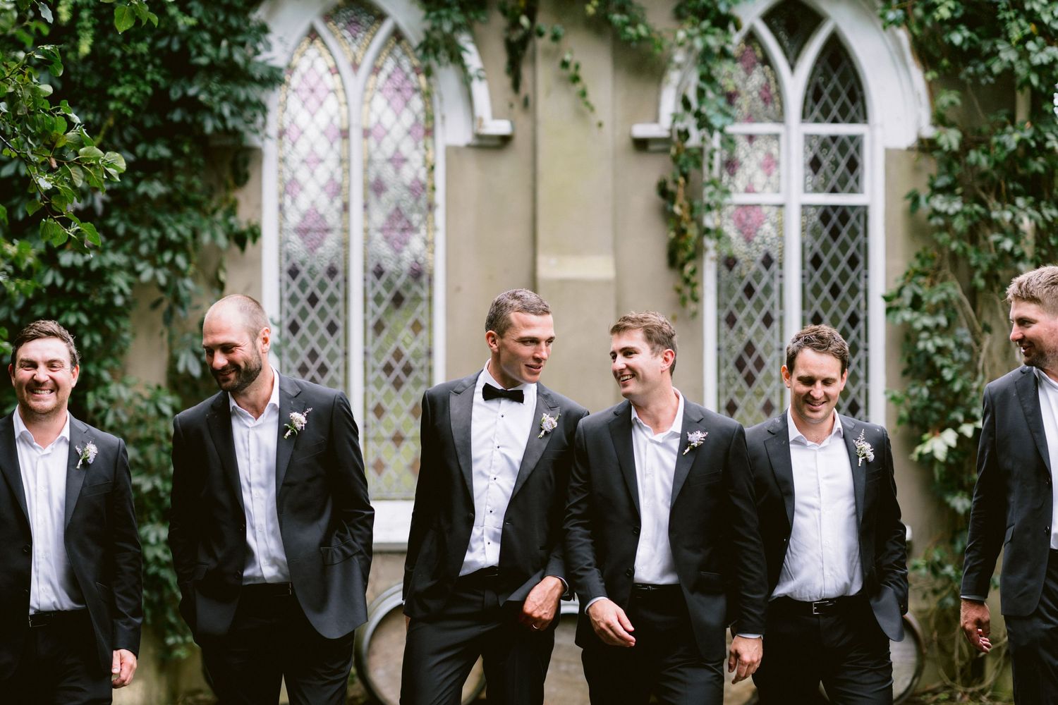 A group in formal black tie attire stands together against an ivy-covered stone building.
