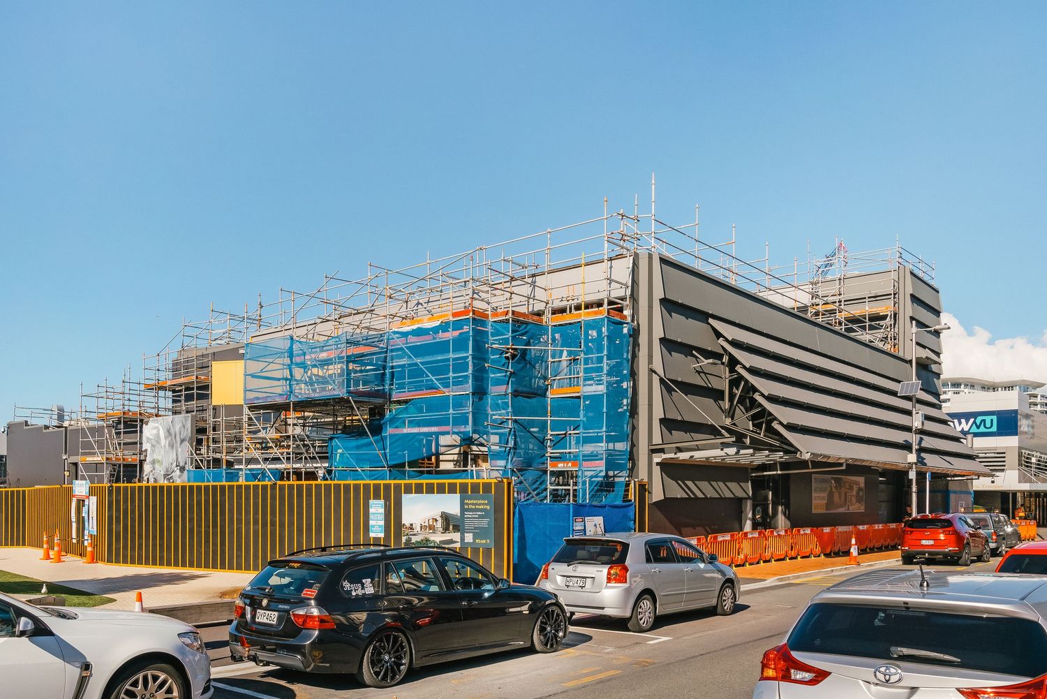 Construction site with multiple commercial vehicles and shipping containers under scaffold-covered buildings on a sunny day.