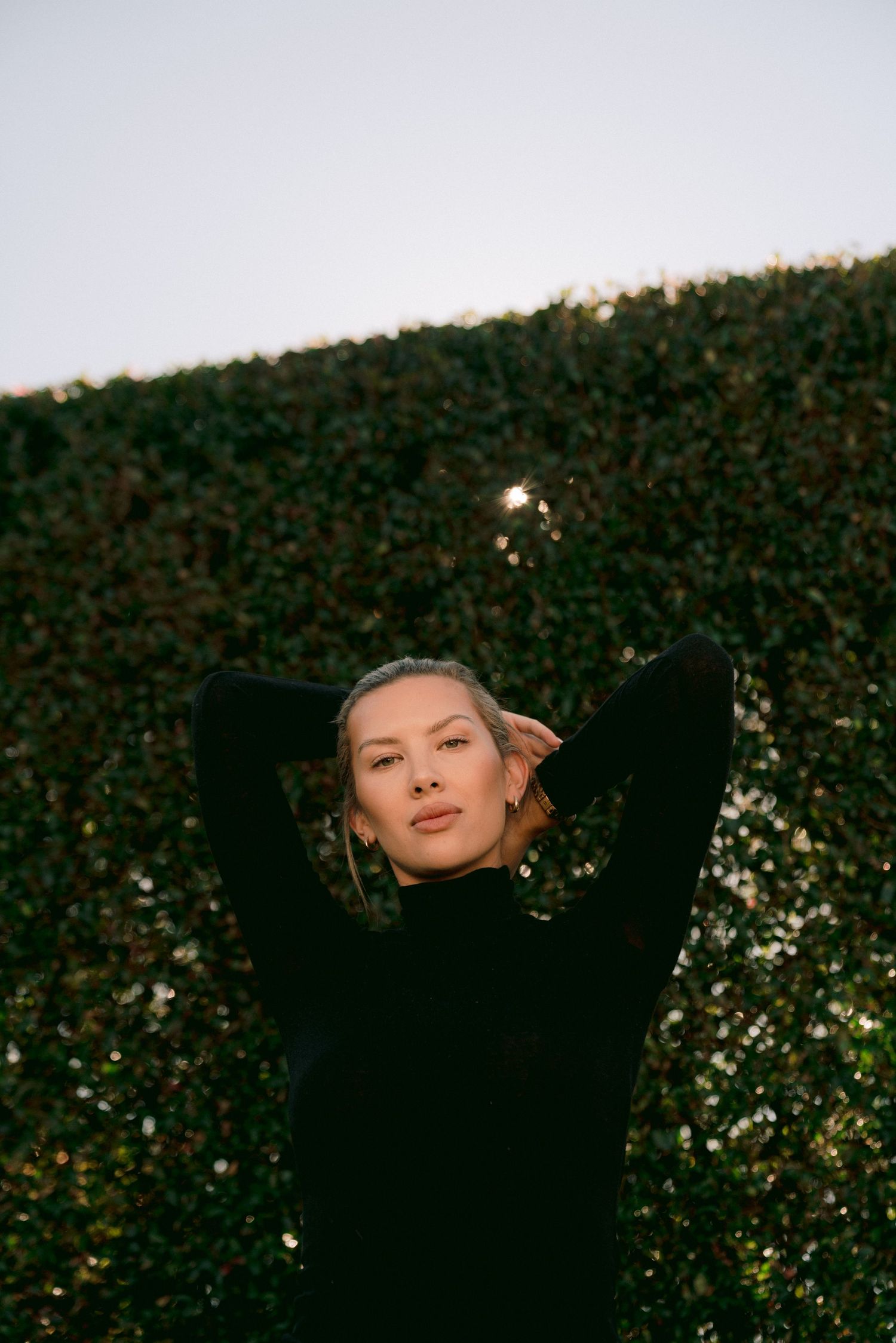 Person in black sweater posing against green hedge wall at dusk in moody outdoor portrait.