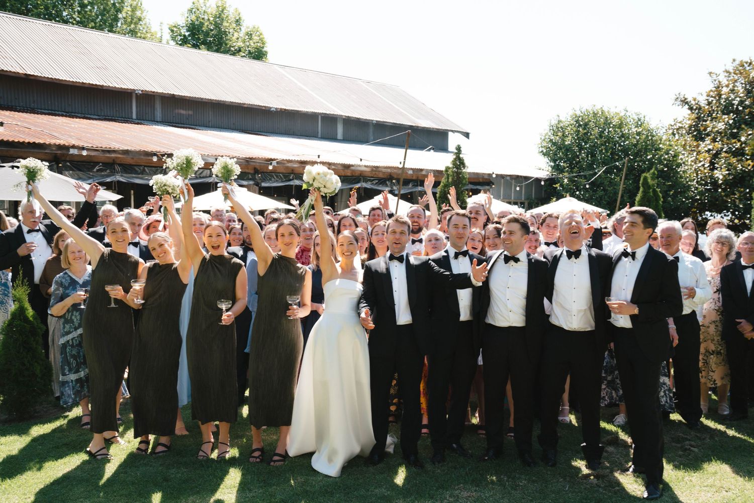 Large outdoor wedding party gathering with guests cheering and celebrating on a sunny day at a venue with green lawn.