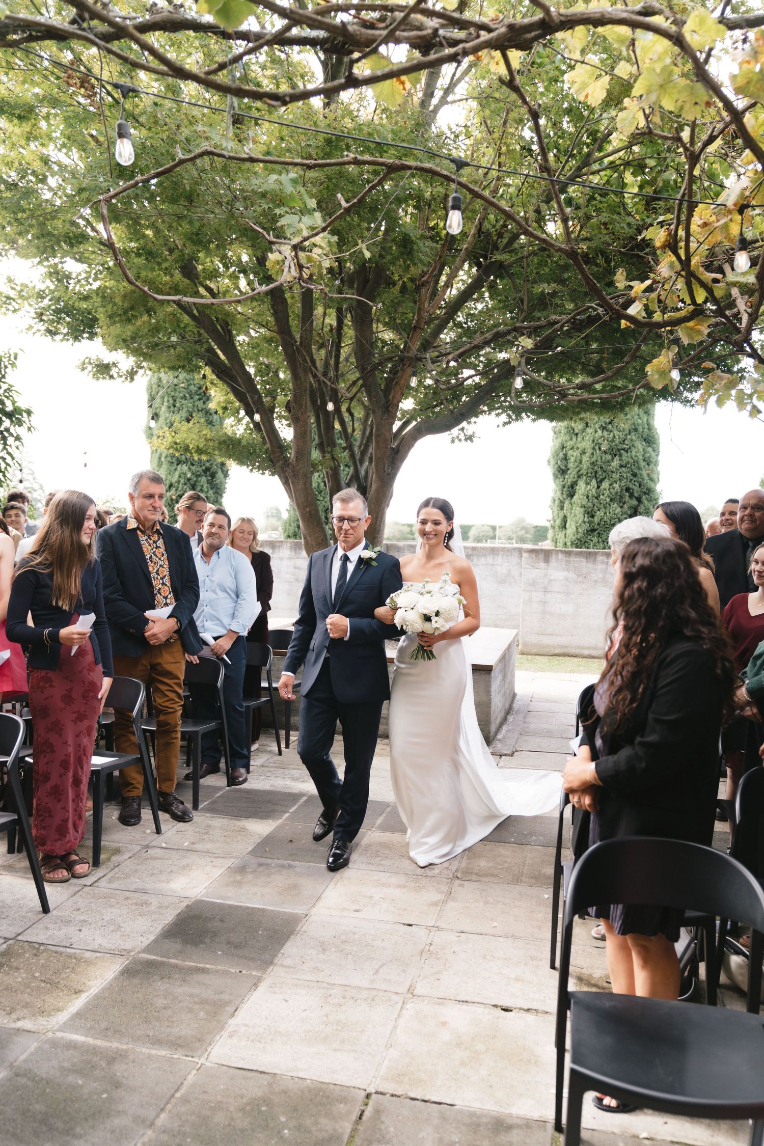 A couple walks down a stone aisle during their outdoor wedding ceremony beneath a canopy of trees.