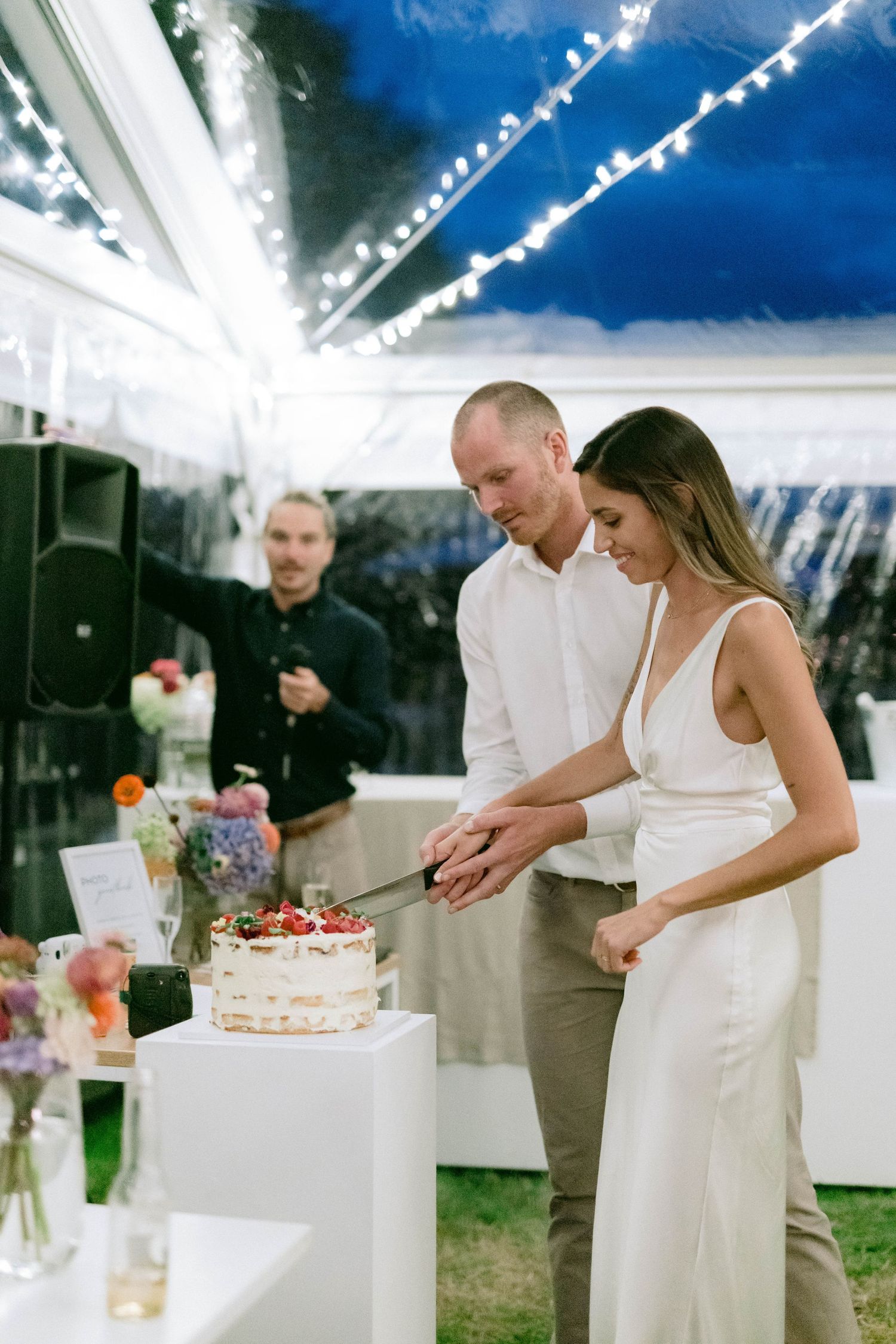 Couple cuts their wedding cake under a tent with string lights during an outdoor reception.