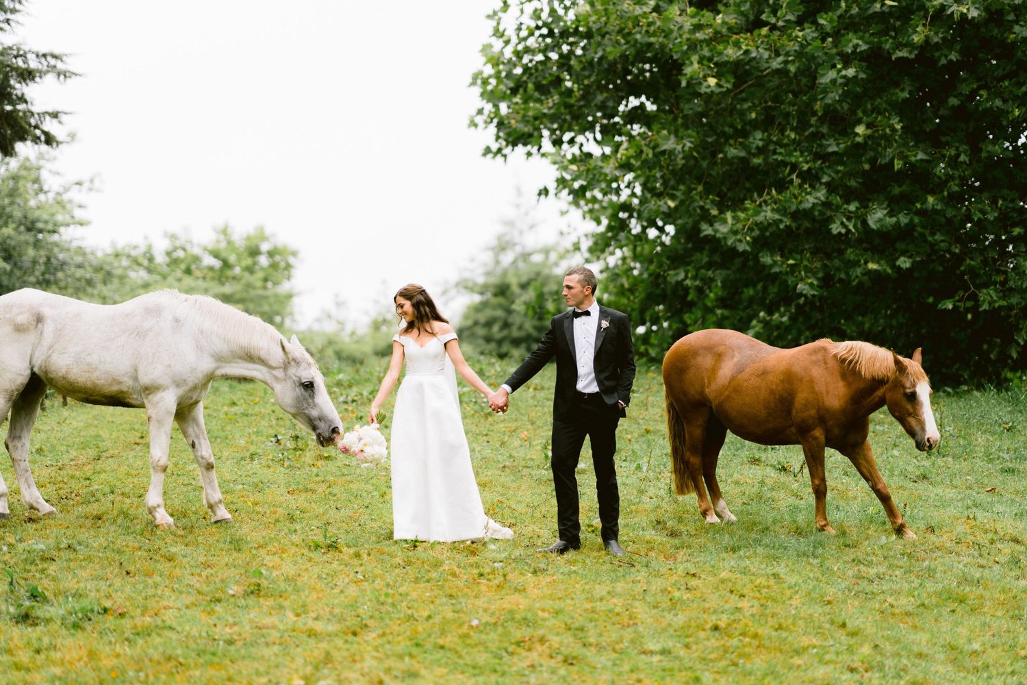 Newlyweds stand between a white and brown horse in a green field.
