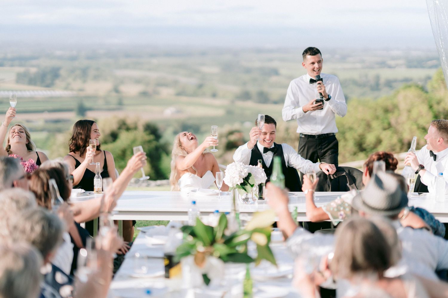 Wedding guests seated at outdoor reception tables raise their glasses in celebration against a scenic mountain backdrop.