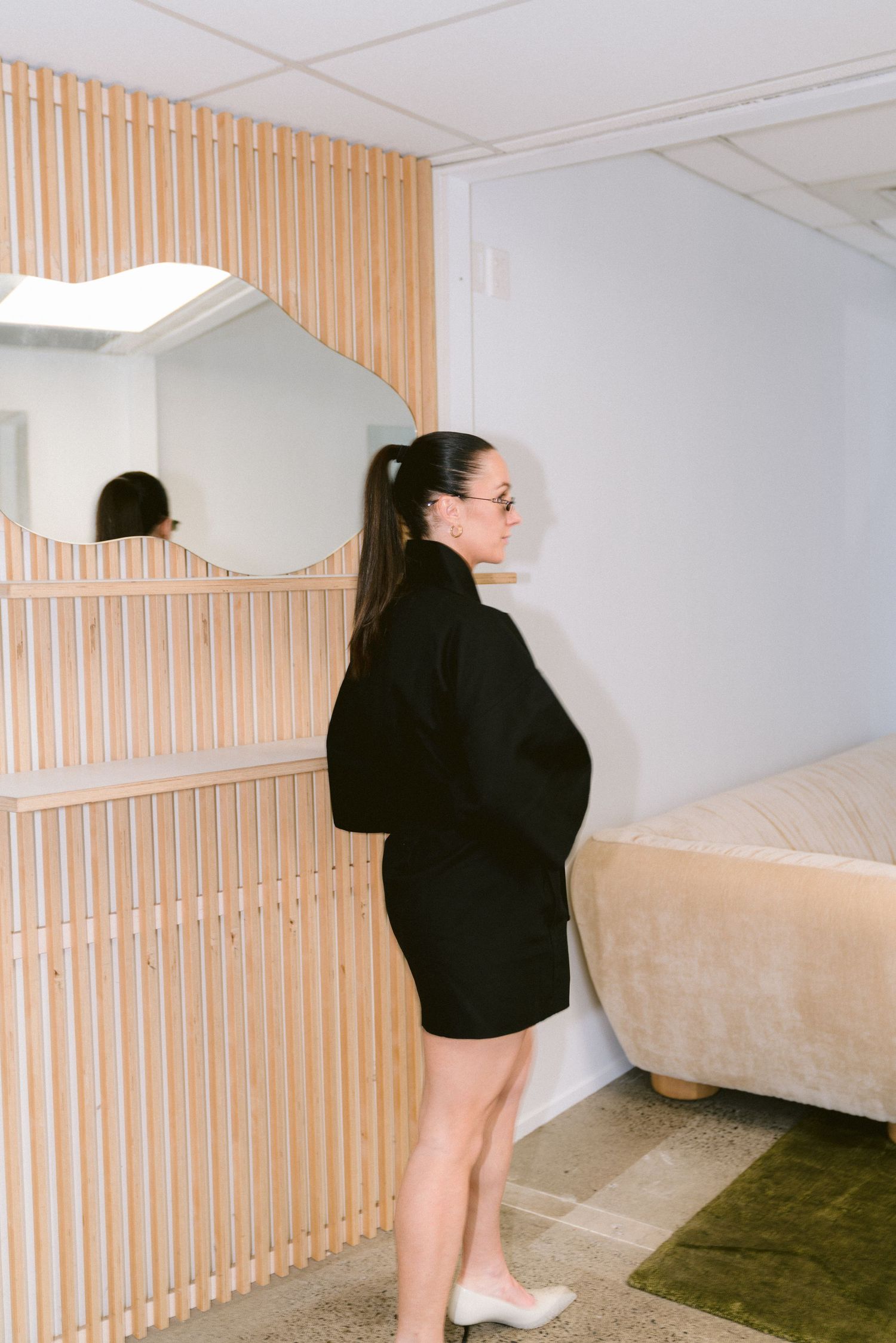 Side profile of someone in a black turtleneck mini dress standing against a wall with slatted wood paneling and curved mirror.
