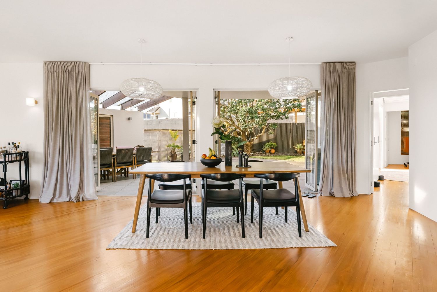 Modern dining room with wooden table, grey chairs, and floor-to-ceiling curtains in bright natural light.