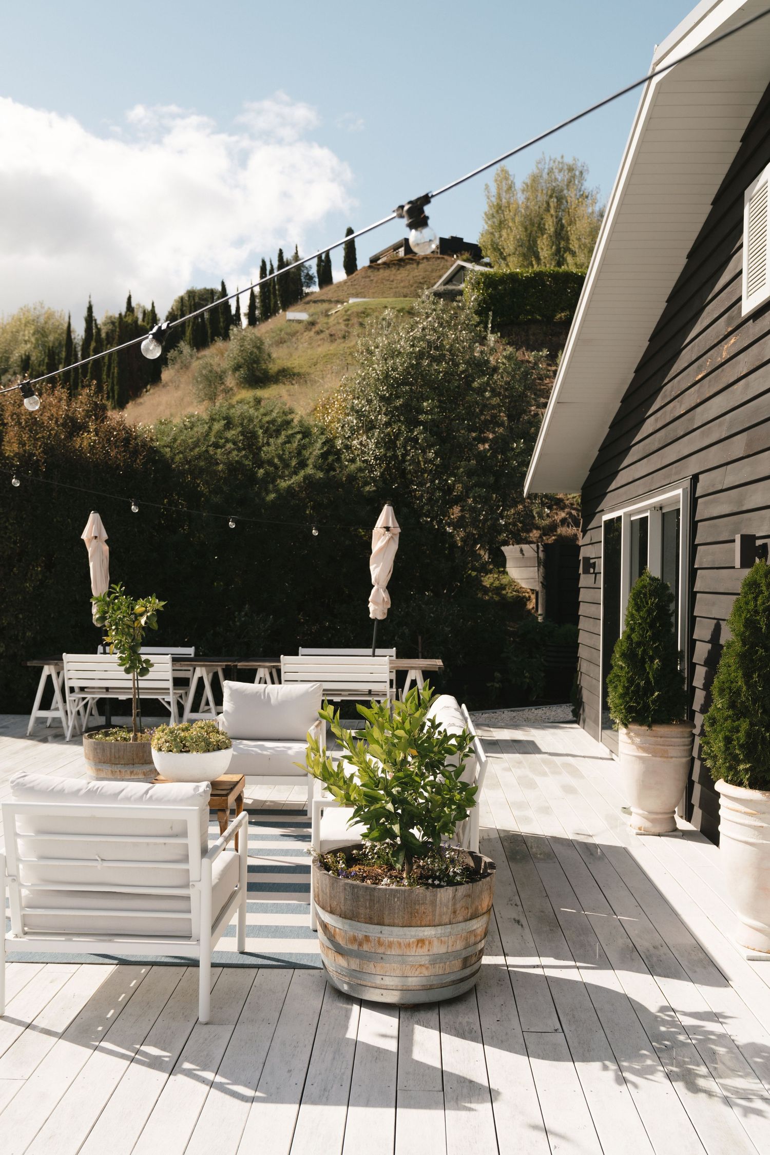 Modern outdoor deck with white furniture and wine barrel planters against a hillside backdrop.