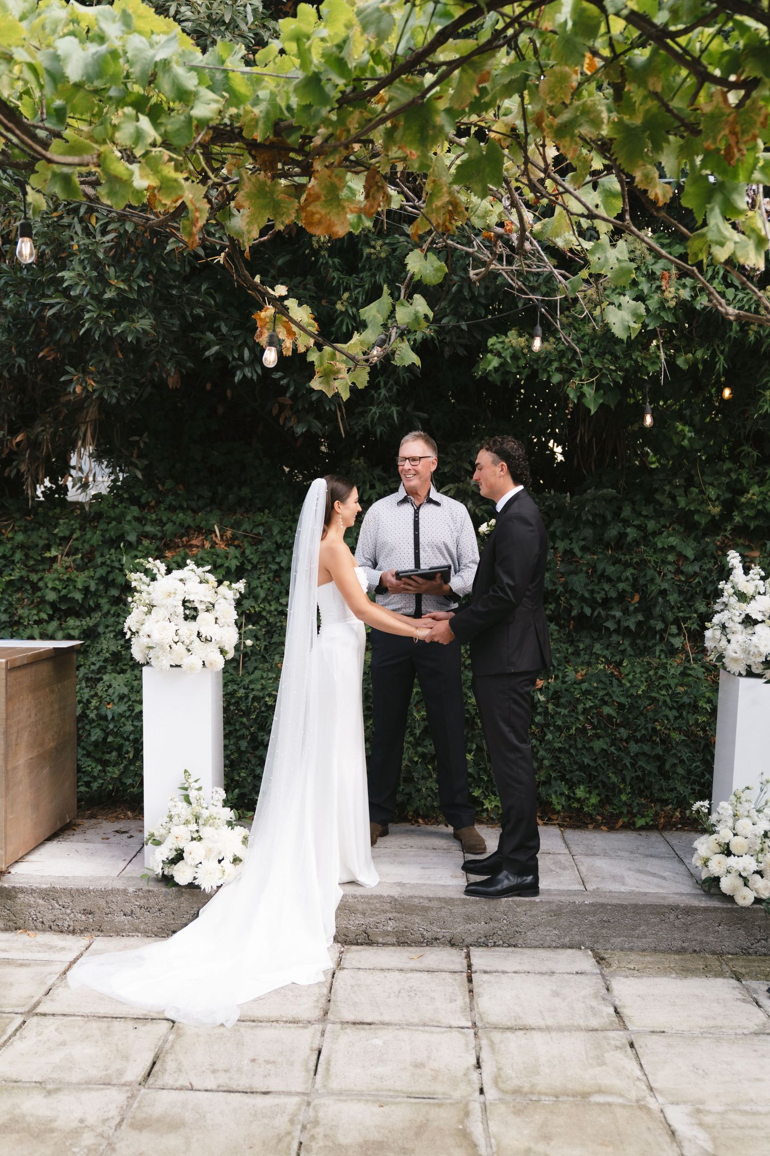 A romantic wedding ceremony takes place in a garden setting with white floral arrangements and greenery overhead.