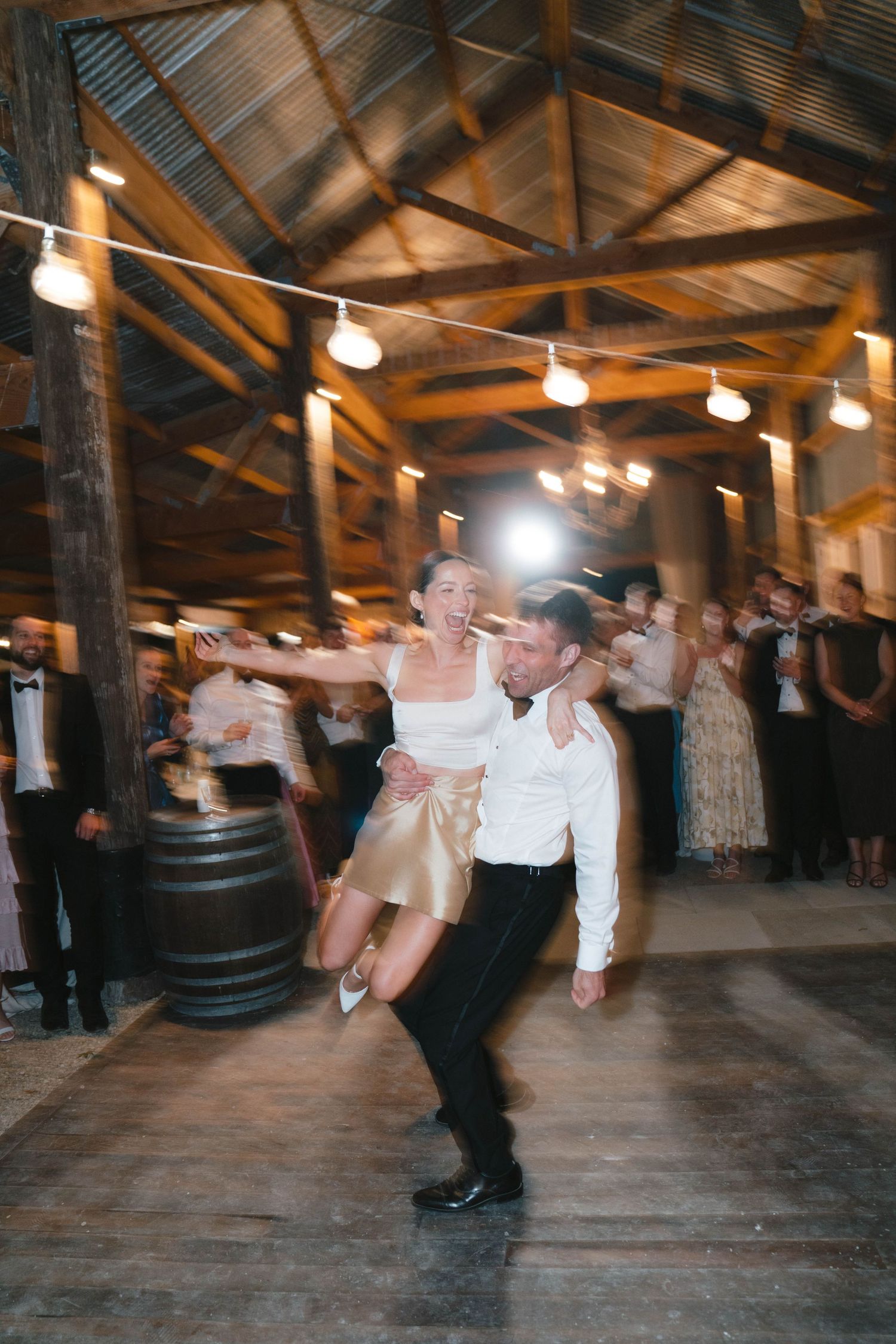 Couple performs energetic dance move during wedding reception in rustic barn with string lights.