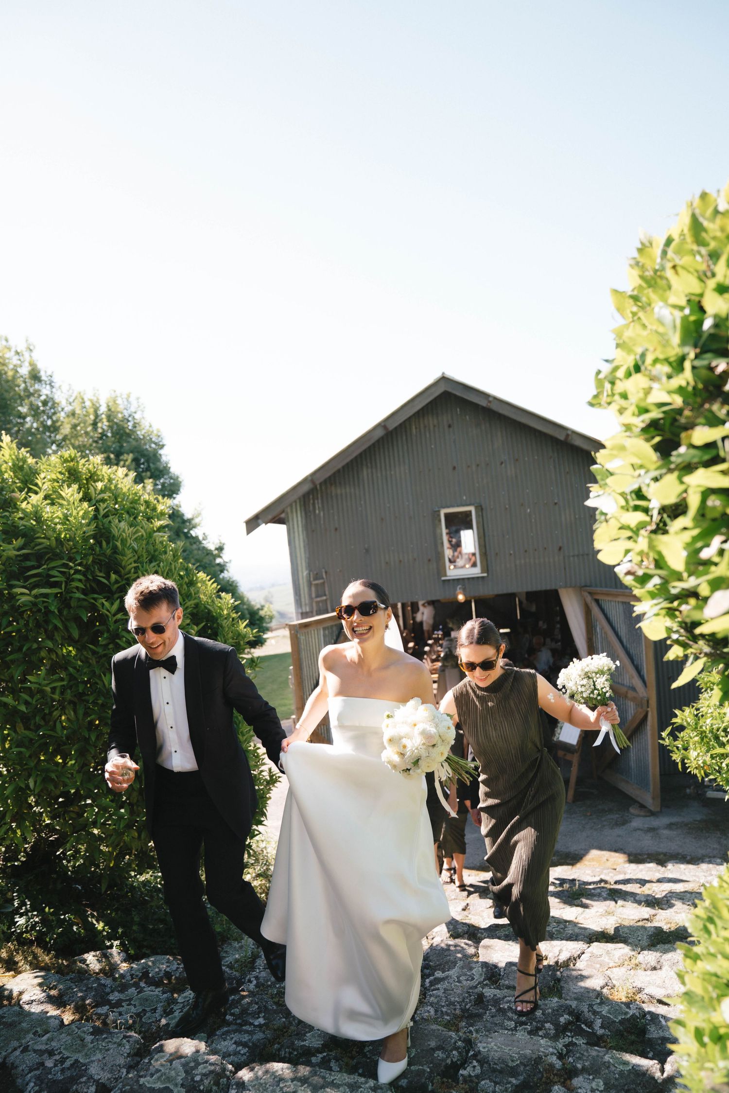 People walking down a rocky path beside a gray barn structure on a sunny day.