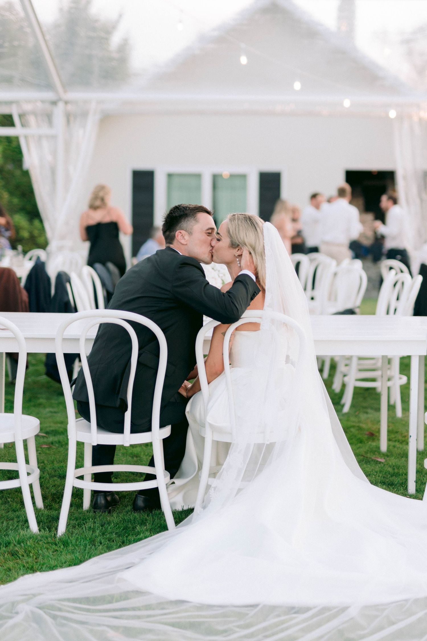 Intimate moment between newlyweds seated on white chairs at outdoor reception.