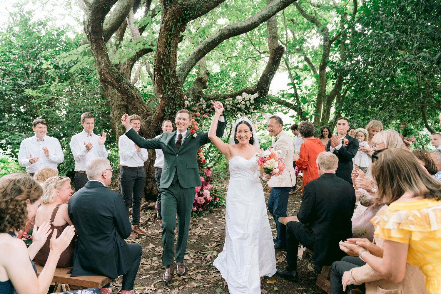 Outdoor wedding celebration under large tree canopy with guests gathered in summer attire.