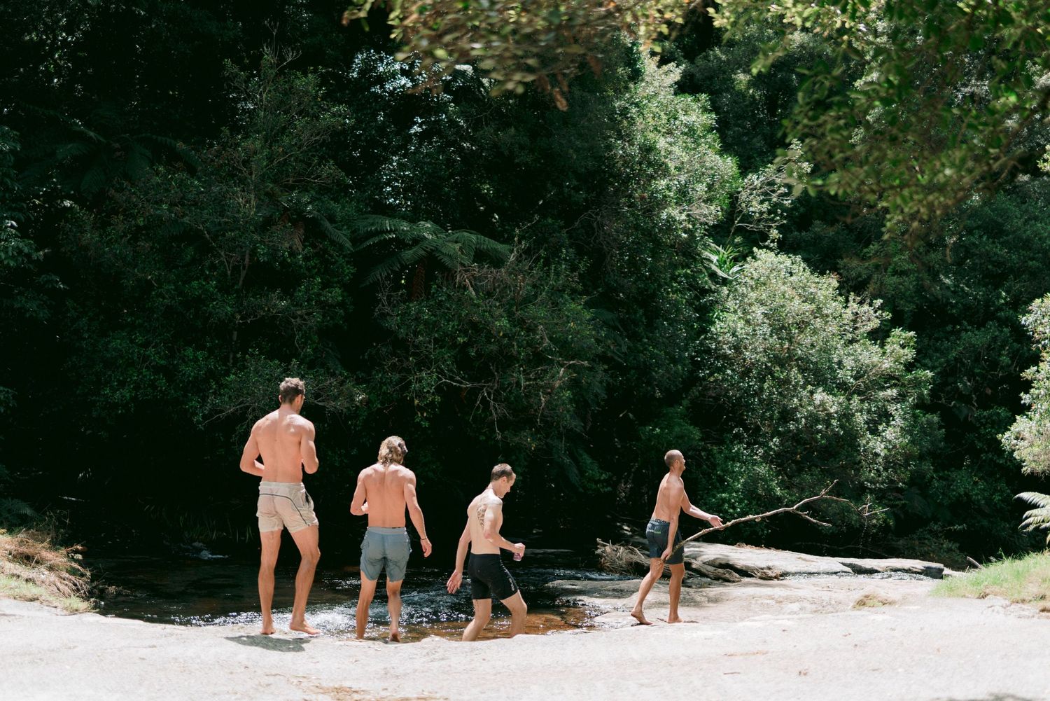 Group walks along sandy riverbank on summer day with lush green trees in background.
