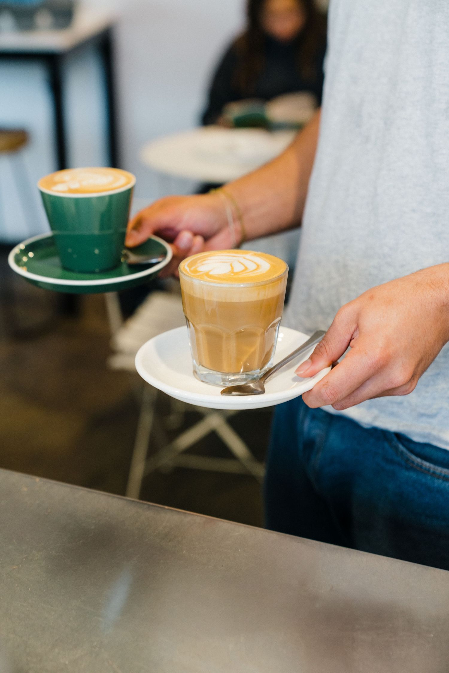 Barista serving two artisanal coffee drinks with latte art in green and glass cups at a modern cafe.