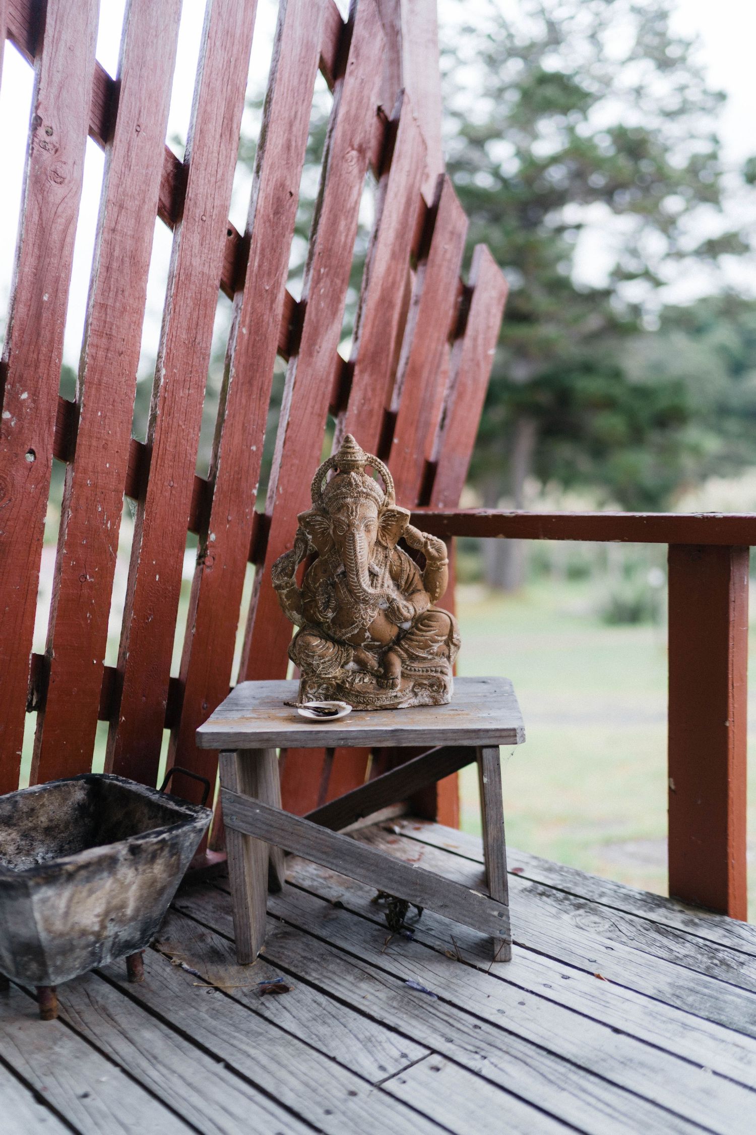 A rustic Ganesha statue sits on a wooden stool against a red slatted fence on an outdoor deck.