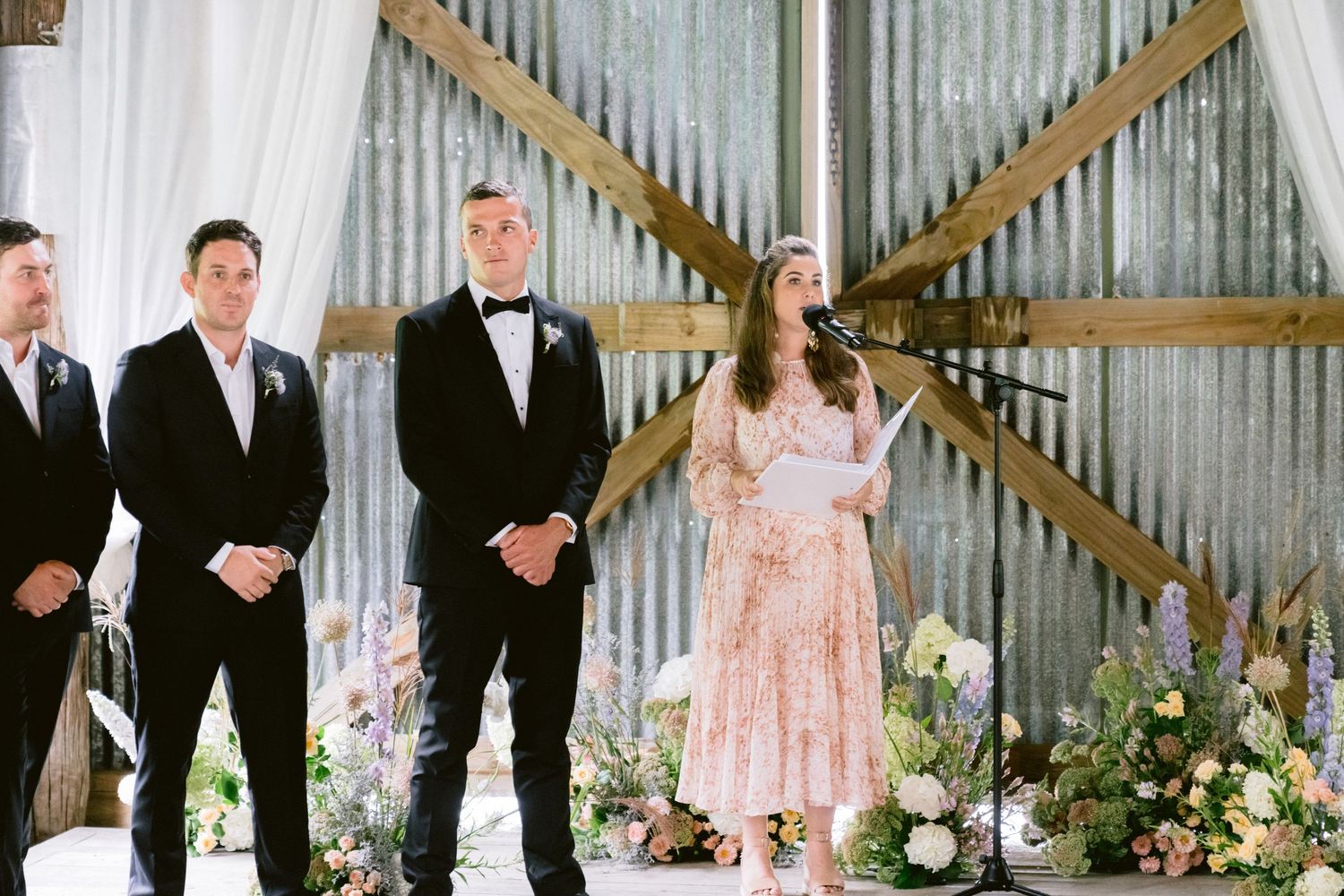 Wedding party poses in rustic wood-paneled barn with elegant floral arrangements and decor.