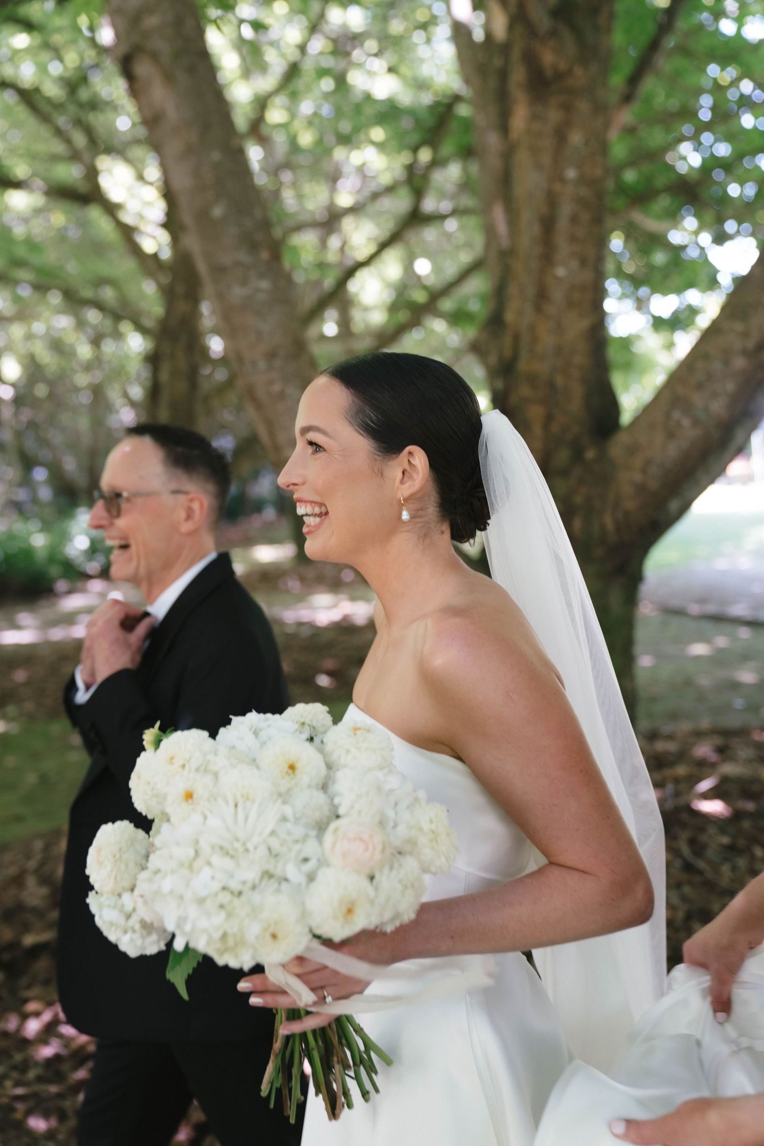 A beaming bride holds a white peony bouquet during an outdoor wedding ceremony.