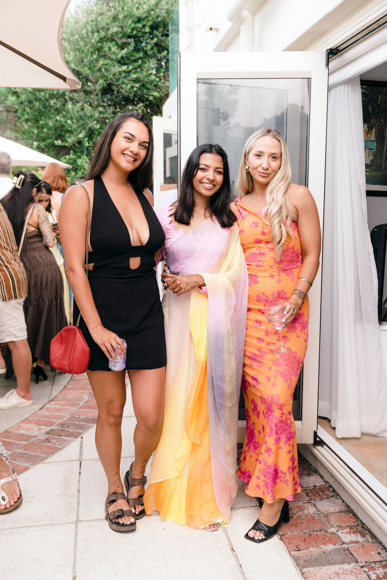 Three friends pose together at an outdoor event wearing fashionable dresses in black, pastel, and vibrant orange tones.