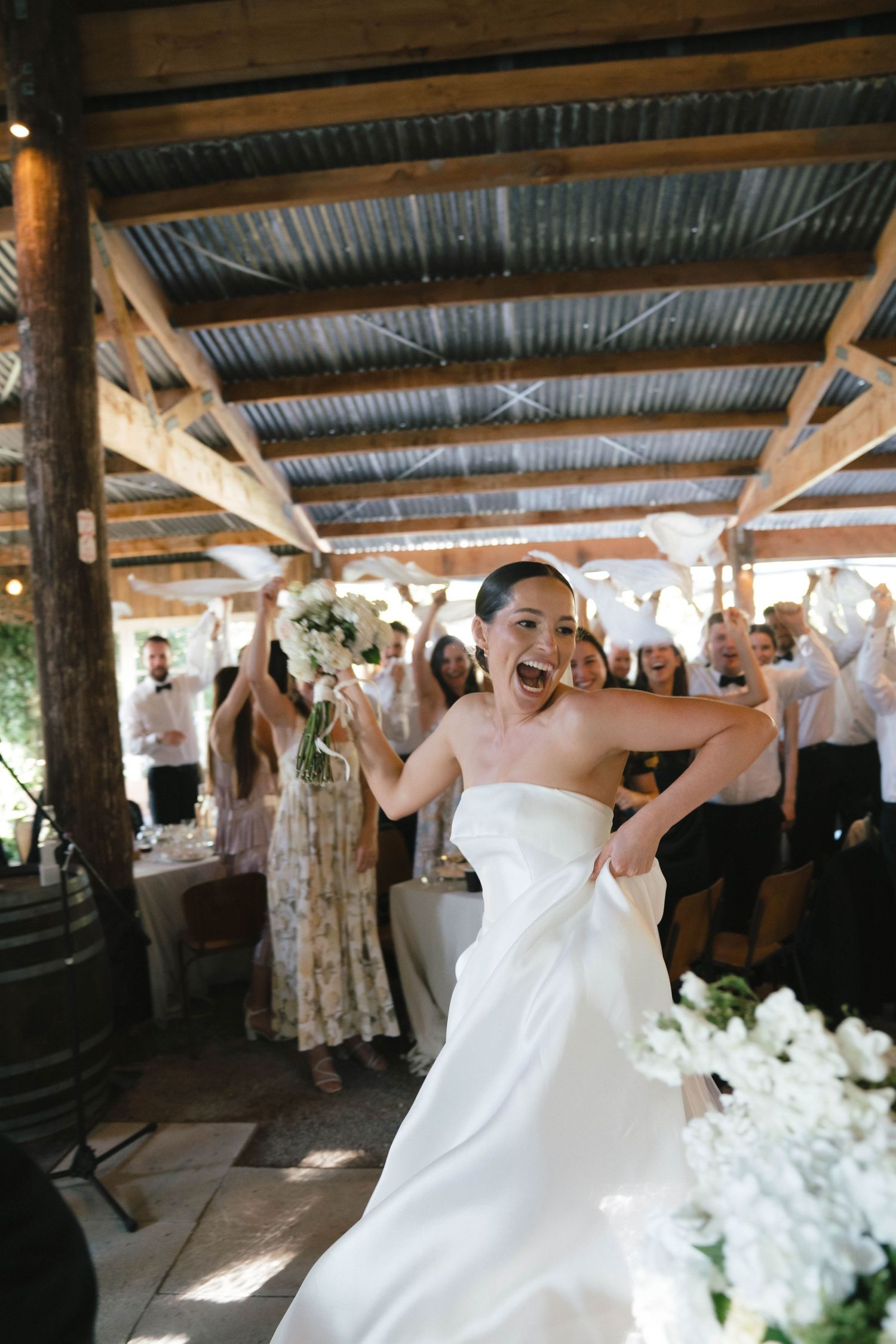 A person in a white wedding dress dances joyfully under a rustic covered venue with guests looking on.