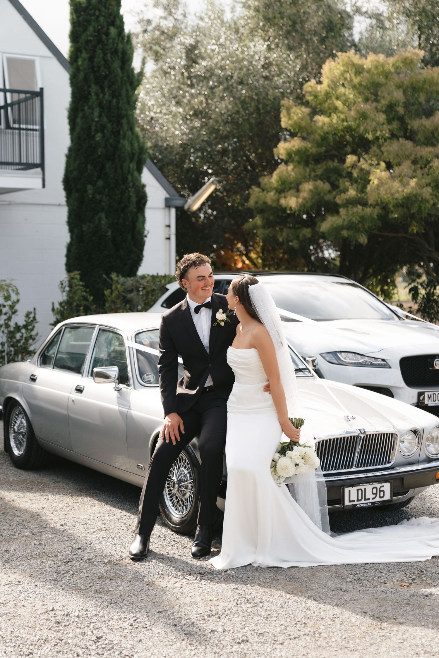 Newlyweds pose playfully beside a vintage silver car after their wedding ceremony.