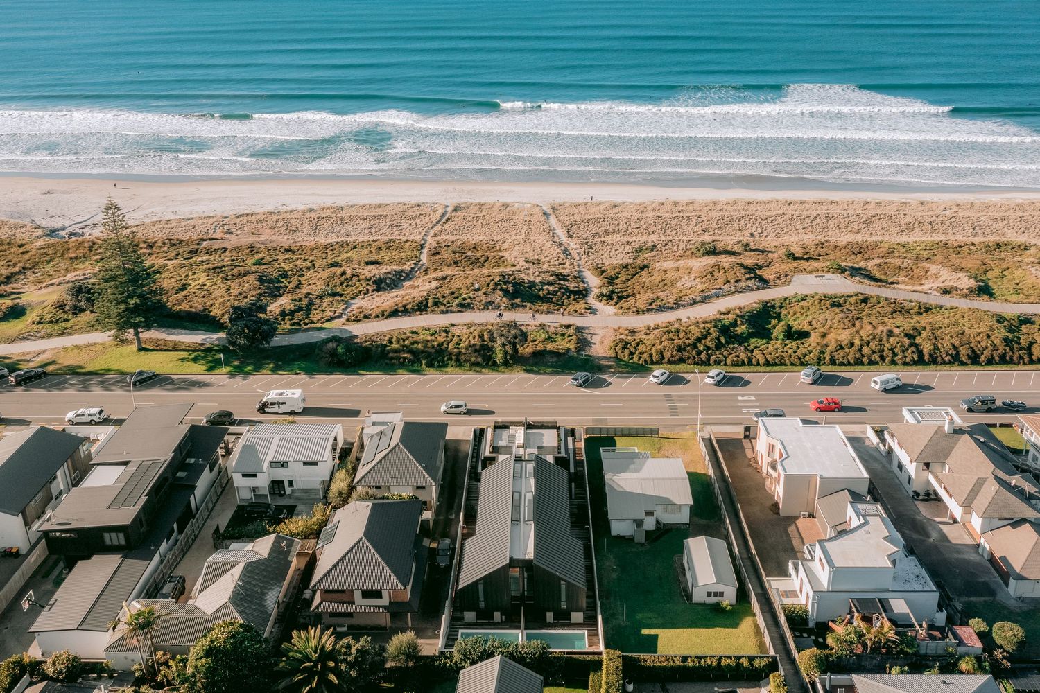 Aerial view of beachfront properties along coastal road with white sand beach and blue ocean.