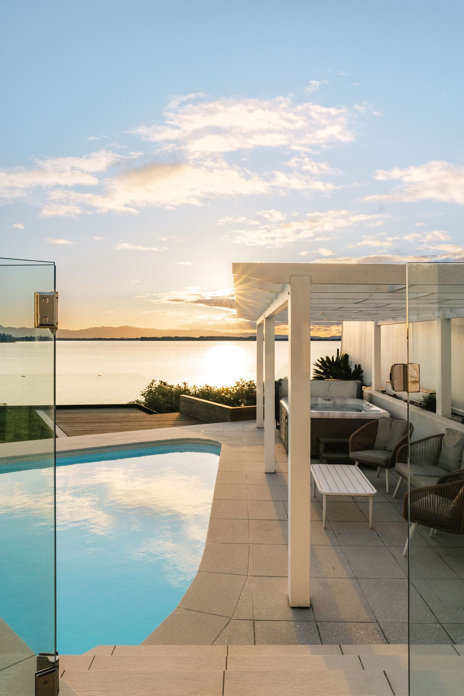 Elegant poolside terrace with glass railings and white columns against a sunset ocean backdrop.