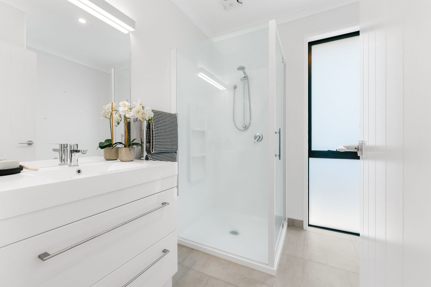 Modern white bathroom interior with glass shower enclosure, floating vanity, and large window providing natural light.