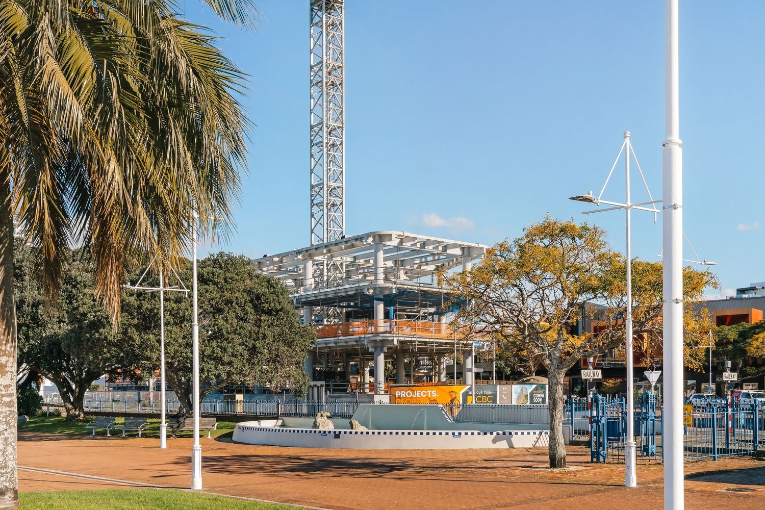 Construction site with palm trees and metal framework visible against blue sky in an urban setting.