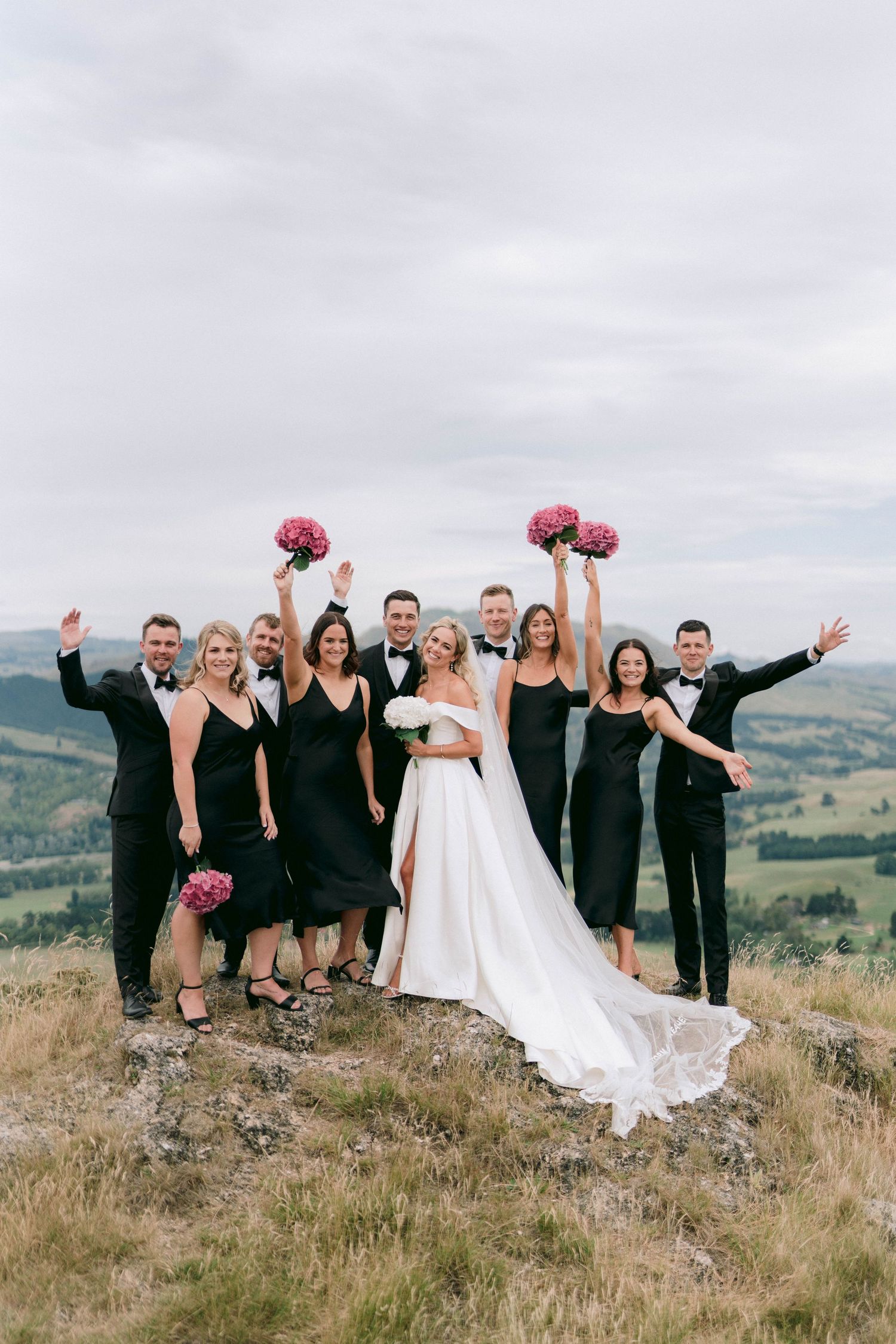 Wedding party in black attire poses on hilltop with pink bouquets against mountain backdrop.